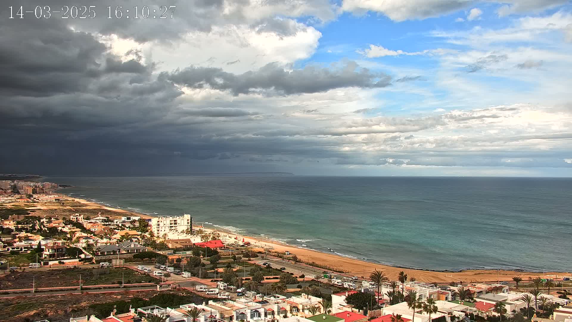 A coastal town with dark, stormy clouds overhead, partially sunny skies, and a wide expanse of ocean with a sandy beach.