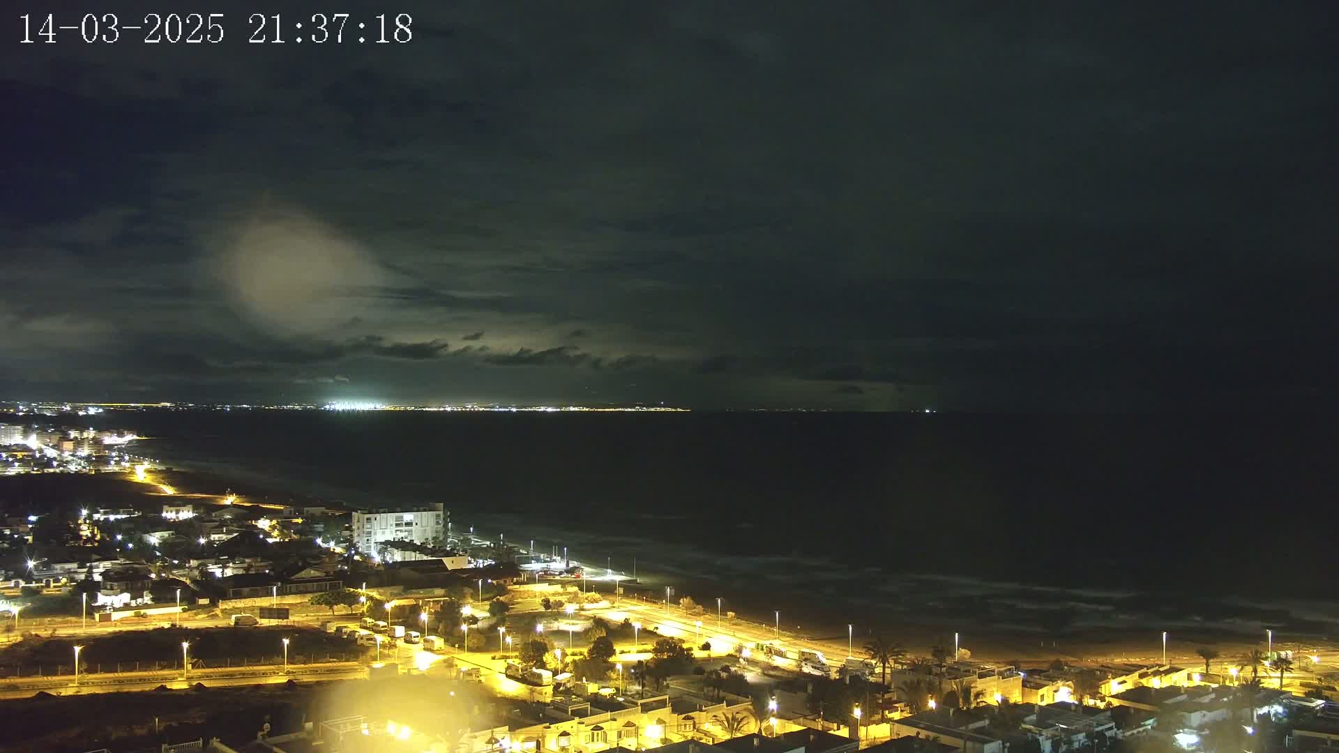 A nighttime coastal scene shows a dark, cloudy sky over a city illuminated with streetlights, a beach, and a dark ocean with small waves.