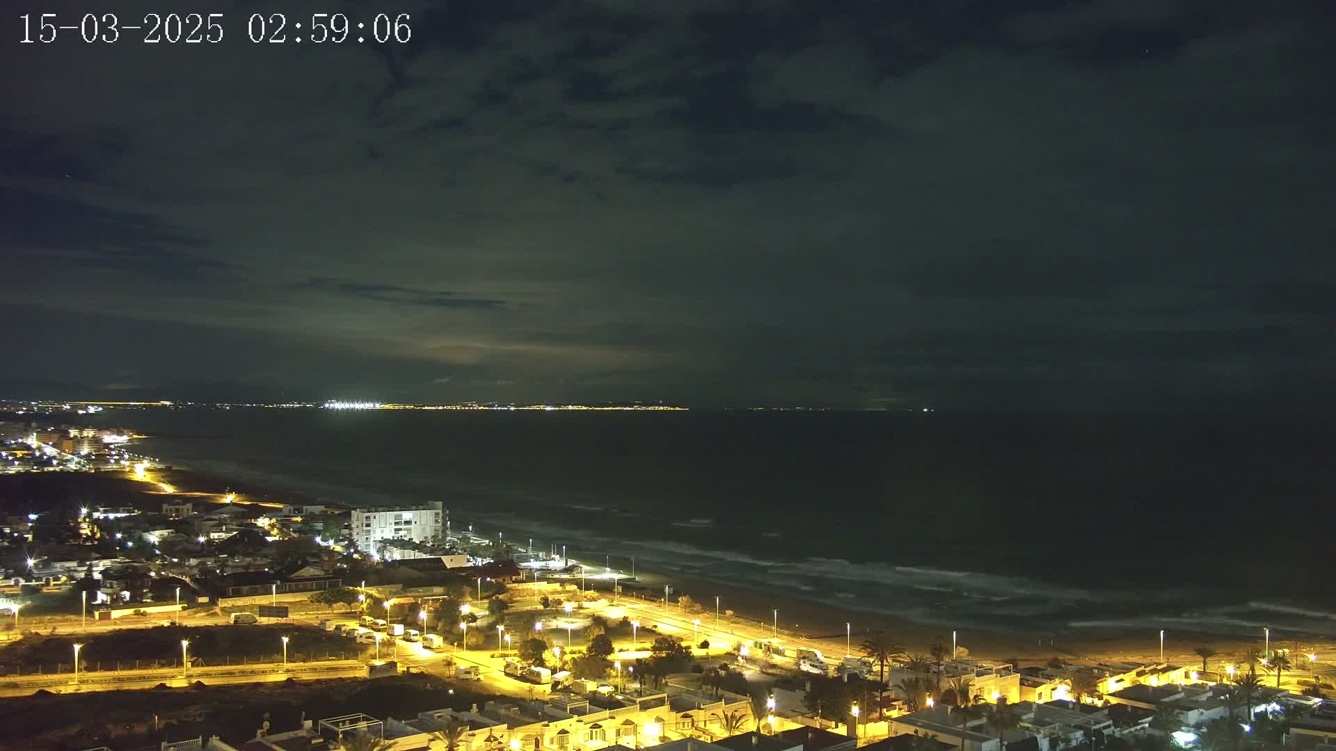 A nighttime coastal scene shows a dark ocean with small waves under a cloudy sky, overlooking a town with illuminated buildings and streets along the shore.