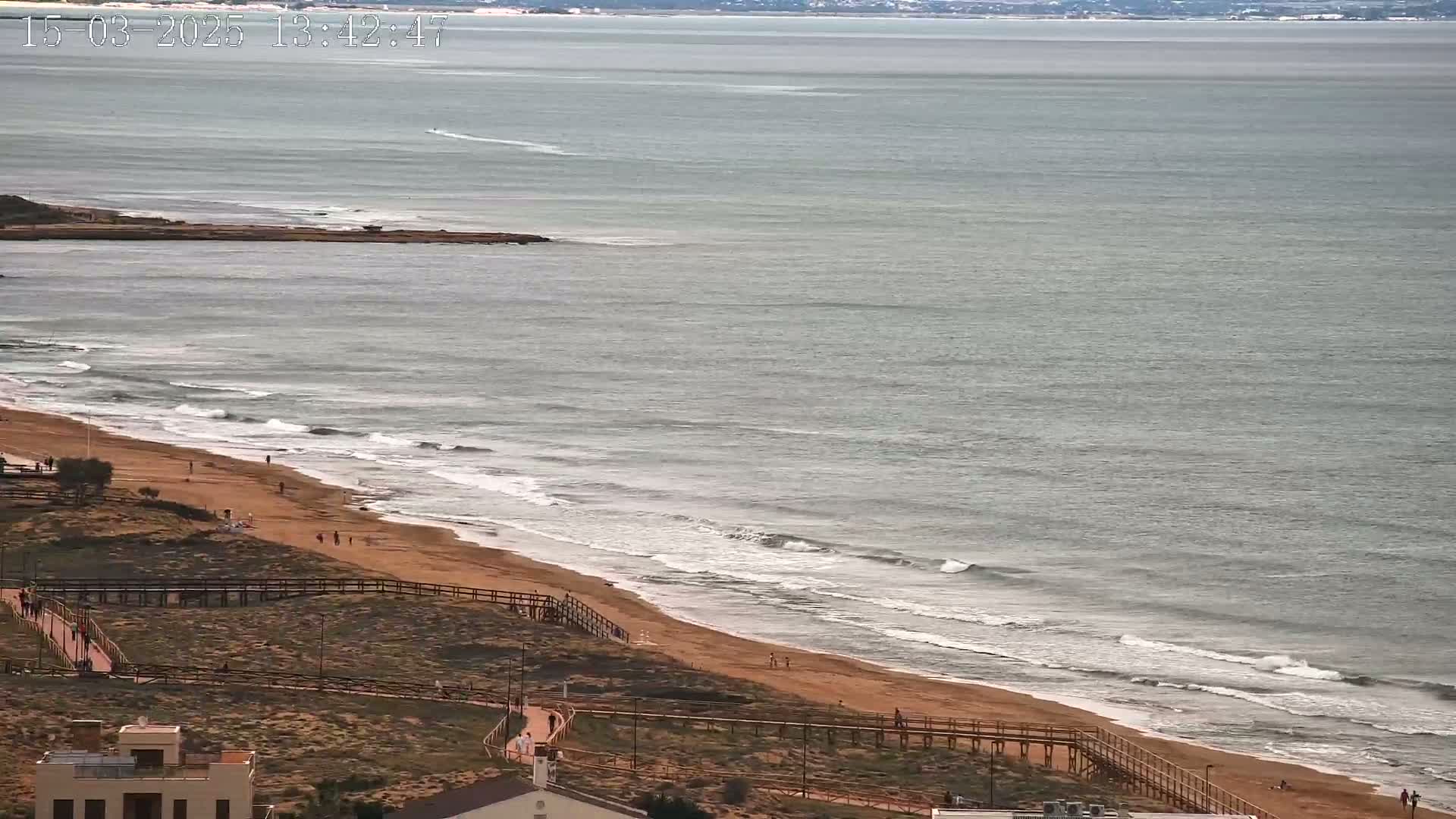 An expansive sandy beach with gentle waves and a few people walking along the shore under a mostly cloudy sky.