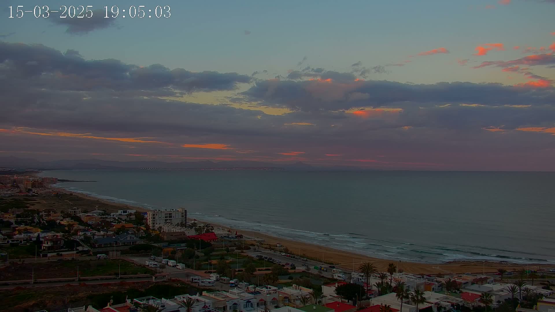 A coastal town at sunset, under a partly cloudy sky with a reddish hue on the horizon, overlooks a calm sea with gentle waves lapping a sandy beach.