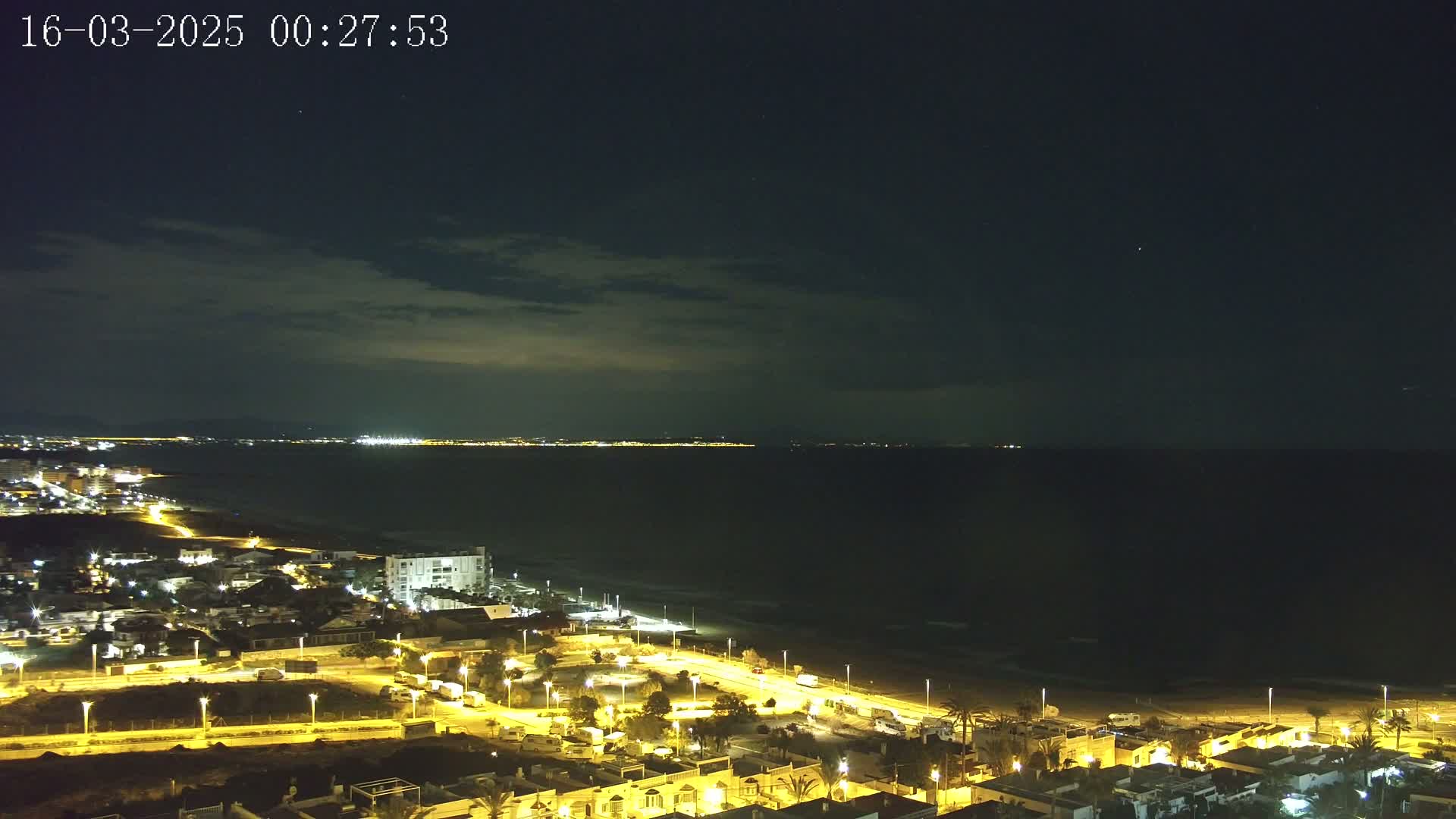 A nighttime coastal scene shows a dark sea under a partly cloudy sky, with a brightly lit town stretching along the beach and distant city lights visible across the water.