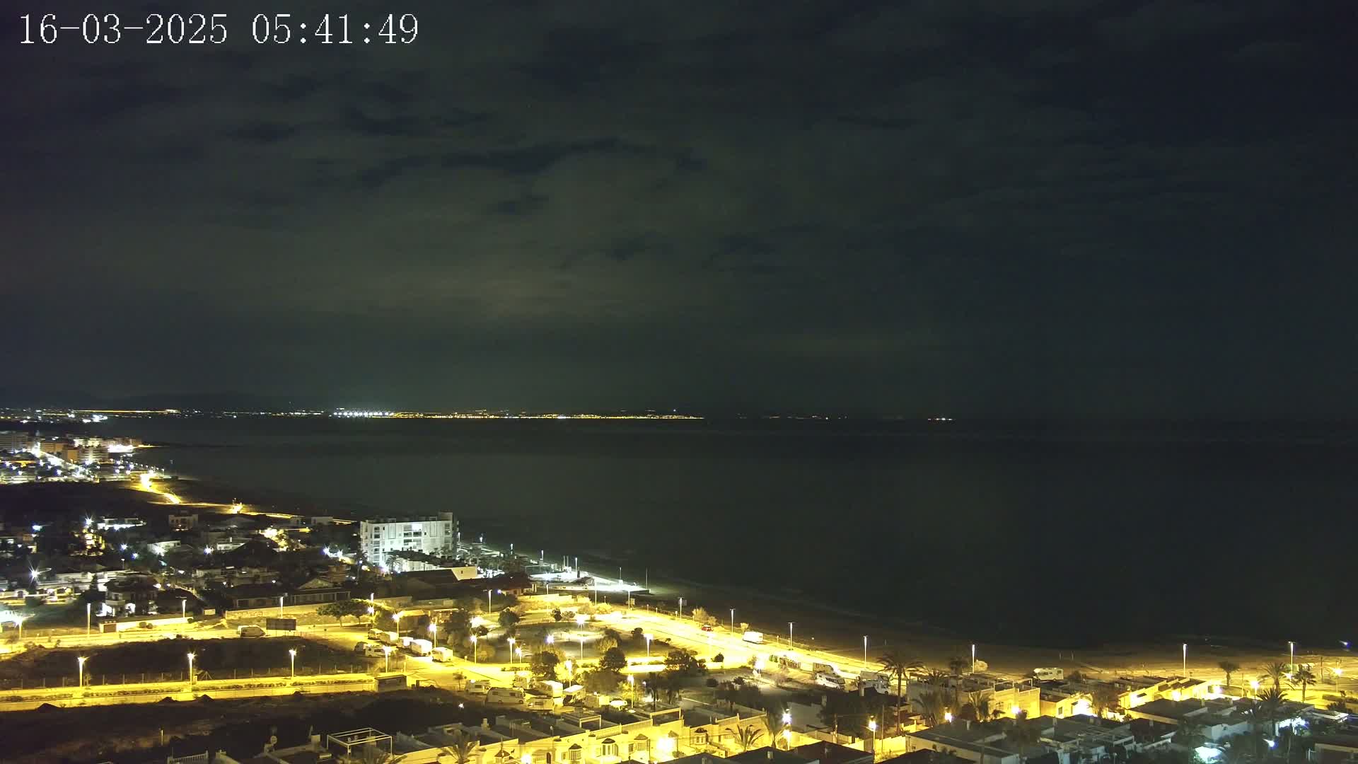 A nighttime coastal town is illuminated, with a dark, cloudy sky above a calm sea.