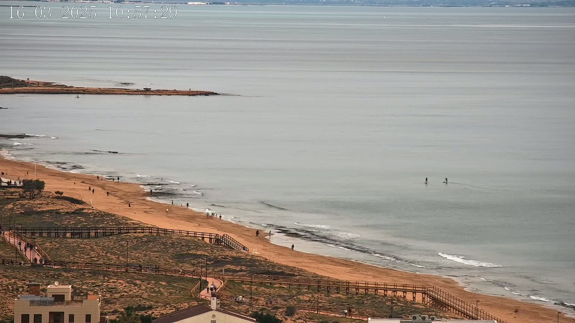 A sandy beach with a boardwalk and scattered people stretches along a calm, grey ocean where two people stand-up paddleboard.