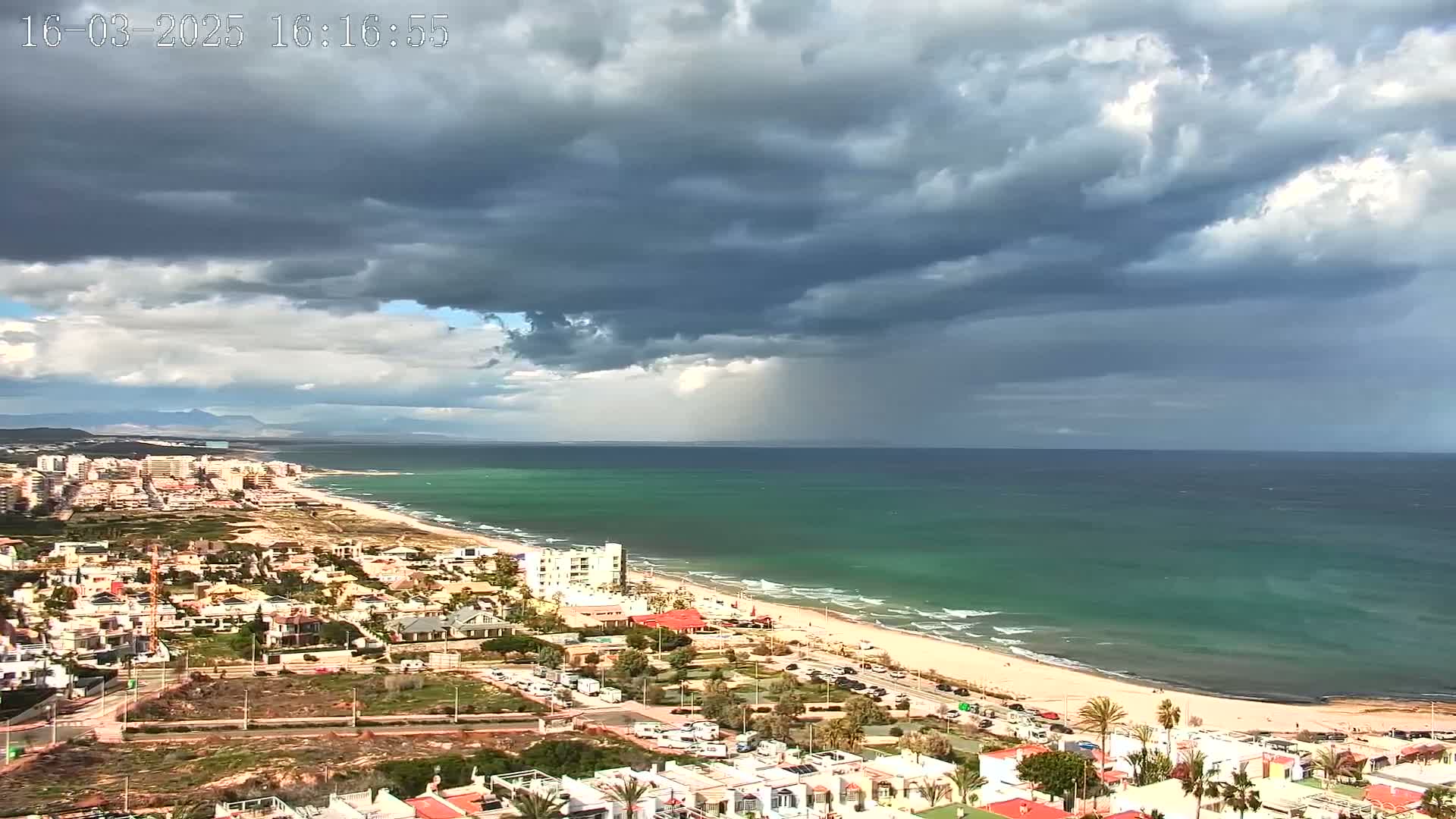 A coastal town with a sandy beach and turquoise ocean is viewed under a dark, stormy sky with rain falling in the distance.