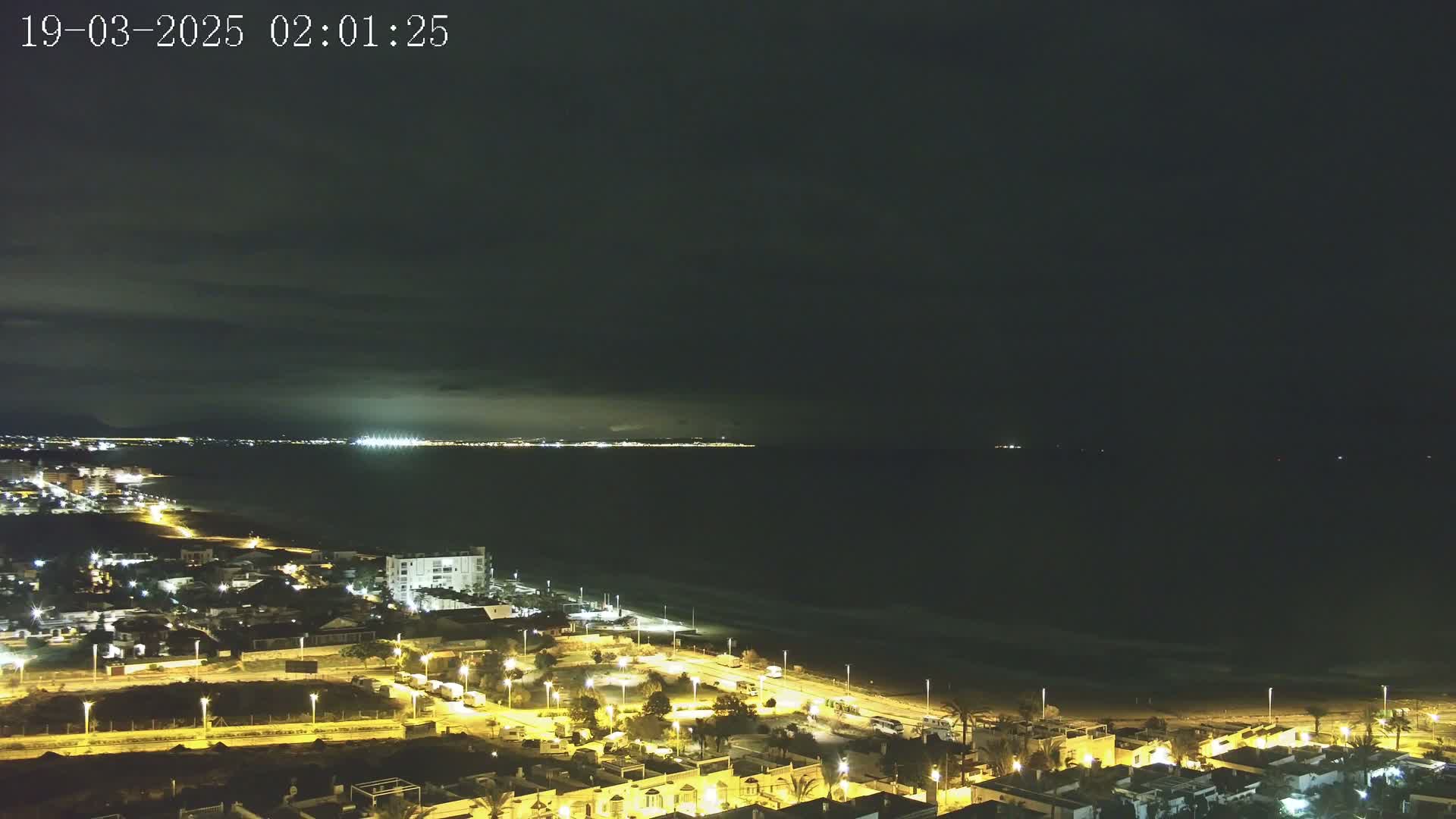 A nighttime coastal town with illuminated streets and buildings is situated next to a dark ocean under a mostly cloudy sky.