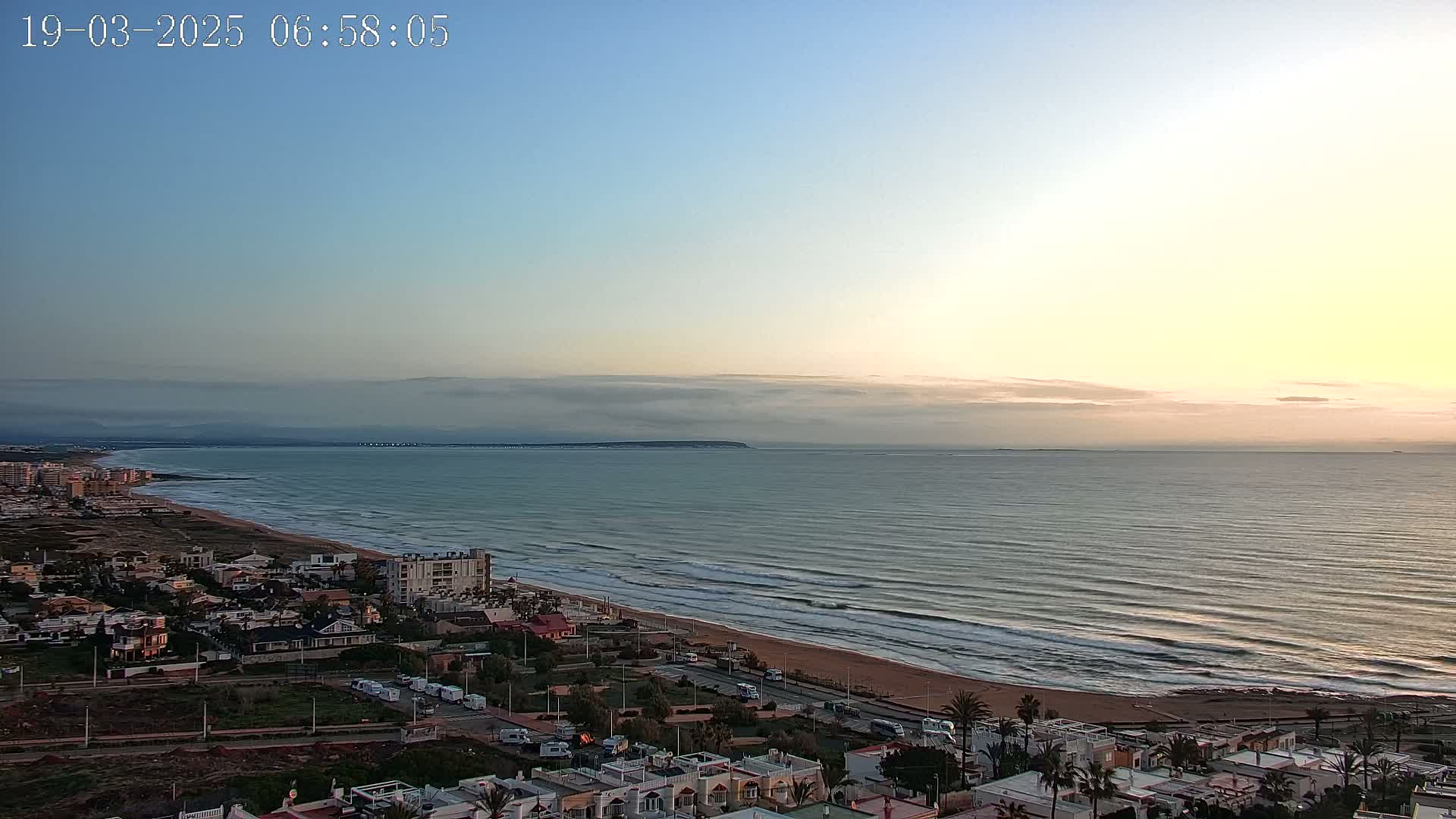 A coastal town at sunrise with a calm sea and a partly cloudy sky.