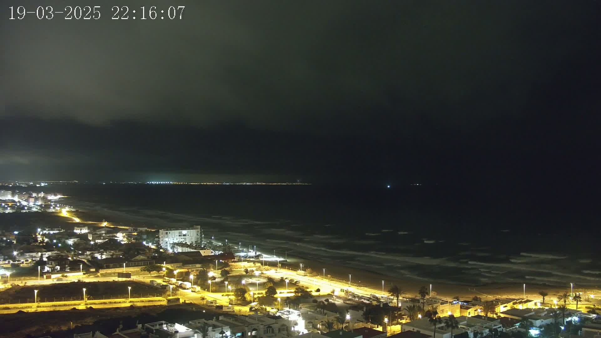 A nighttime coastal town illuminated by streetlights overlooks a dark, wavy ocean under a cloudy sky.