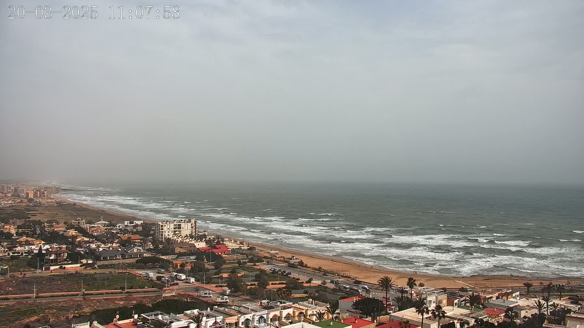 A hazy coastal town overlooks a beach with moderately rough waves under a gray sky.