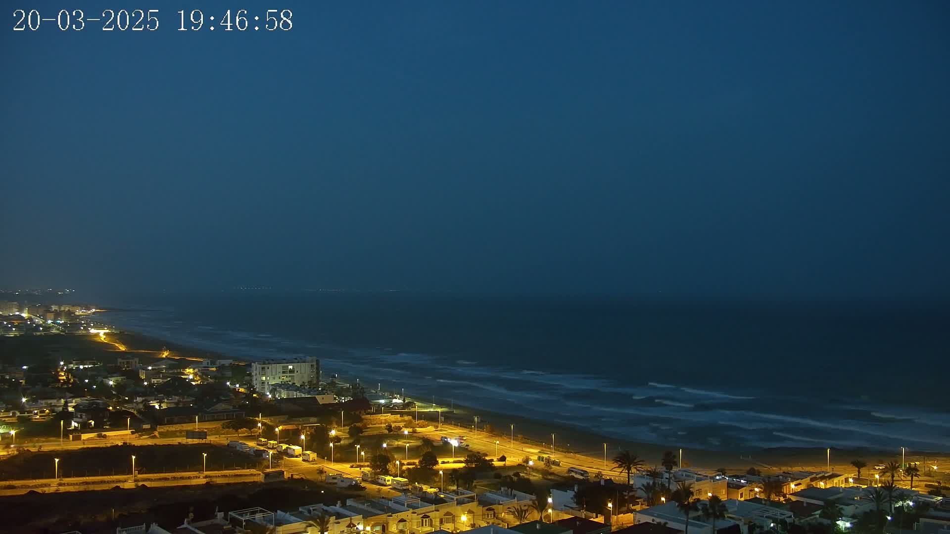 A coastal town at night, under a dark blue twilight sky, with gently rolling waves visible on the ocean.