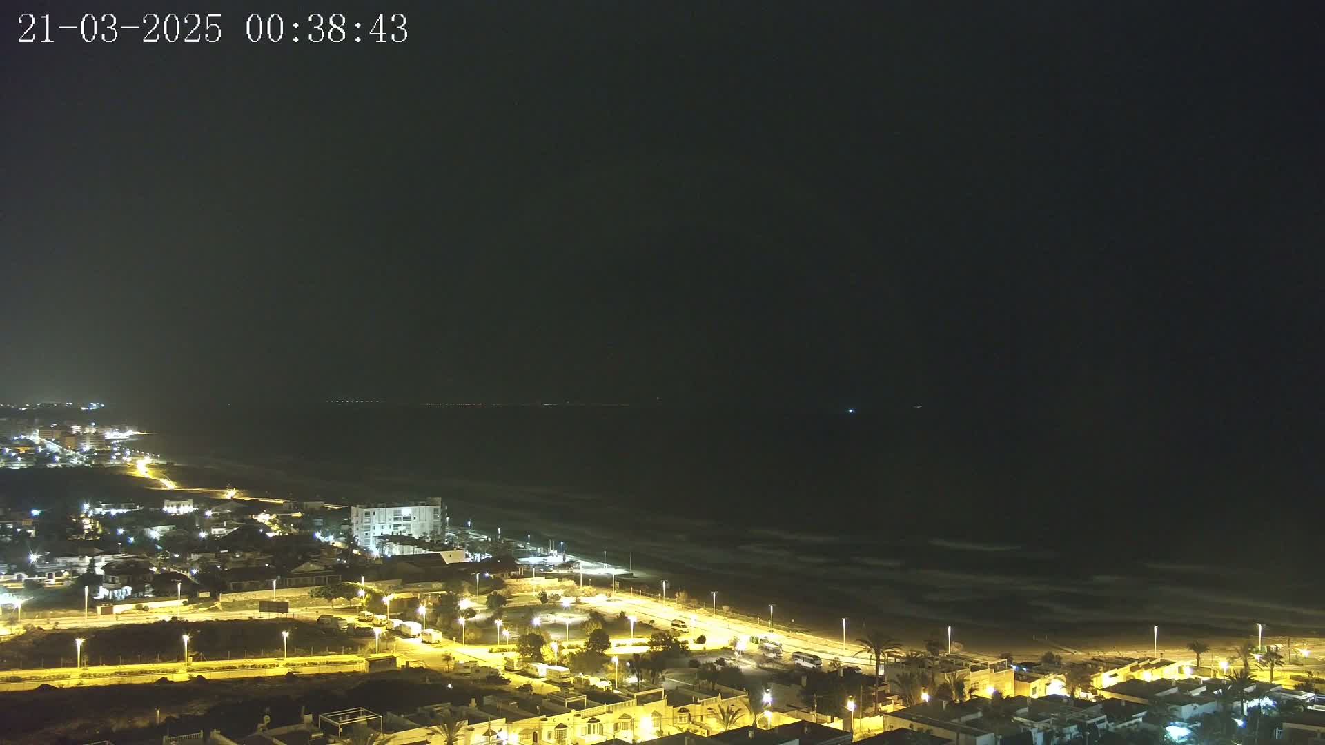 A nighttime view of a coastal town with illuminated streets and buildings overlooking a dark ocean with gently rolling waves under a clear, dark sky.