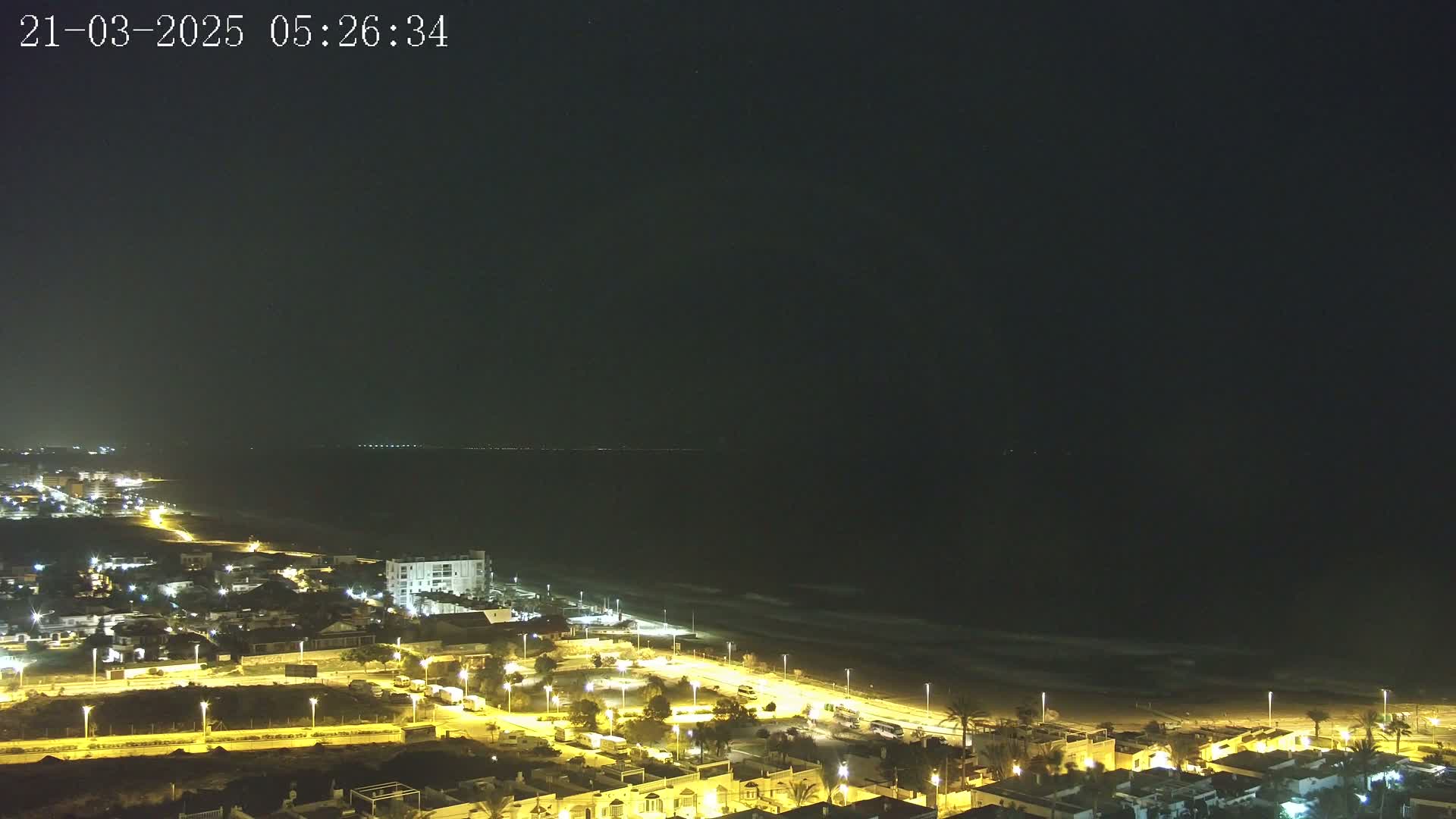 A nighttime coastal scene shows a dark ocean with gentle waves lapping a lit beach, next to a town with illuminated streets and buildings under a clear, dark sky.
