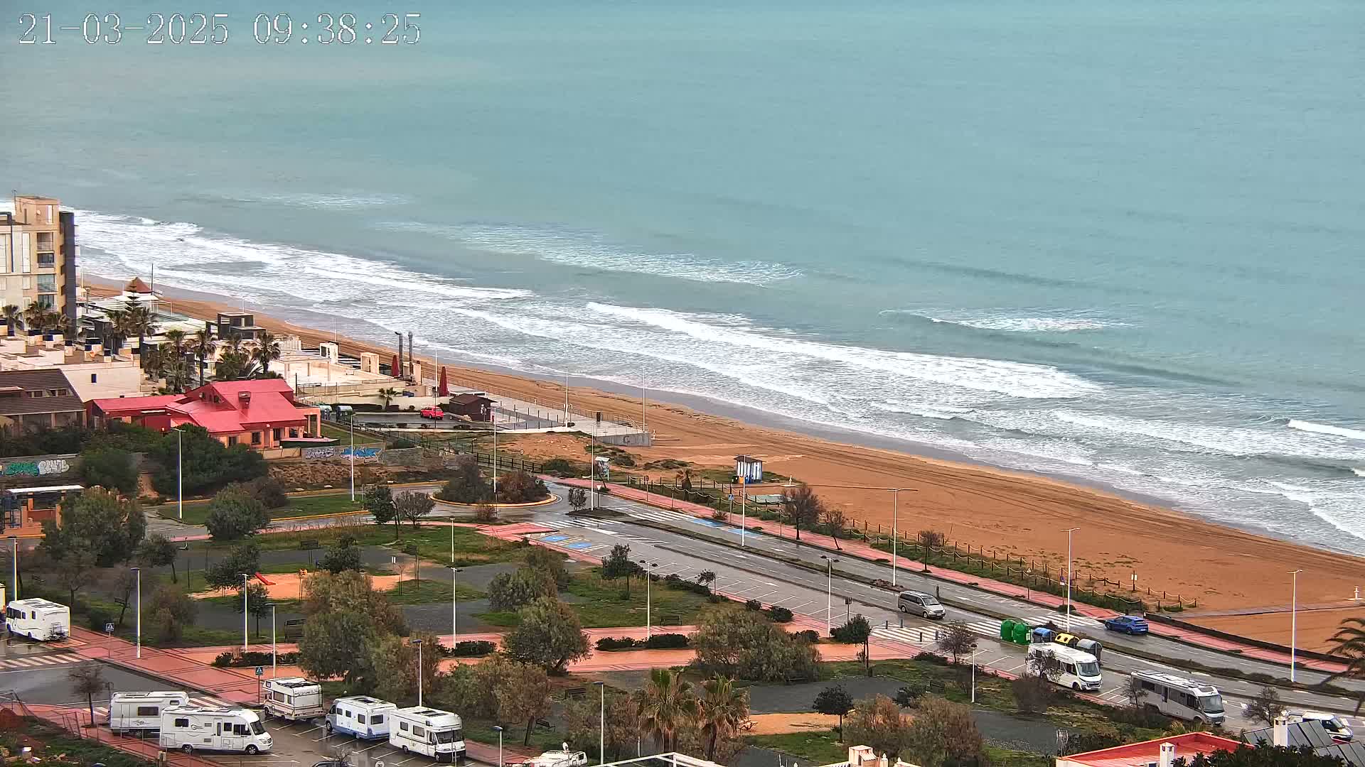An aerial view shows a sandy beach with waves lapping the shore, a road lined with RVs, and buildings nearby on an overcast day.