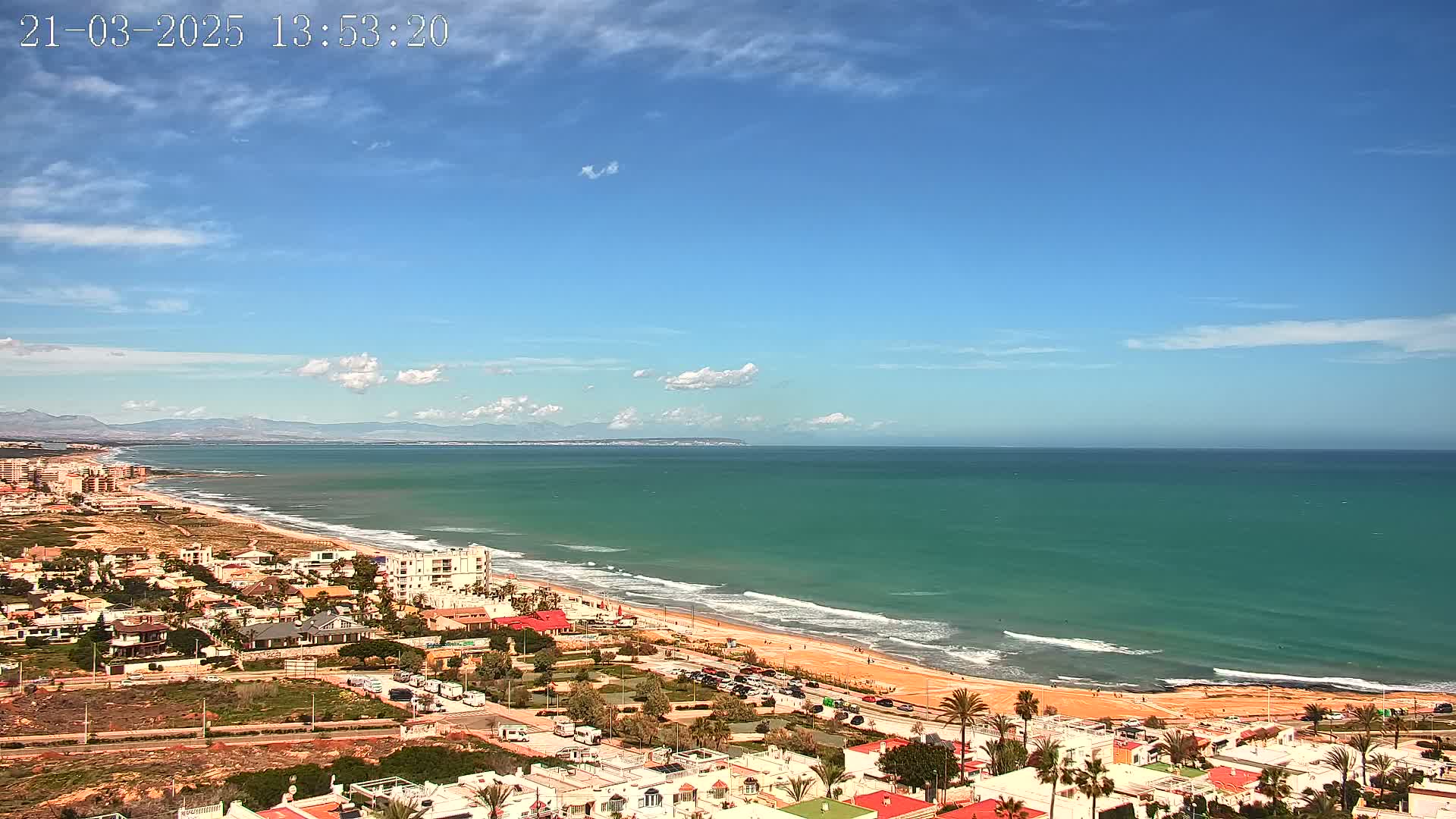 A coastal town overlooks a long sandy beach with turquoise water and small waves under a mostly clear blue sky with a few scattered clouds.