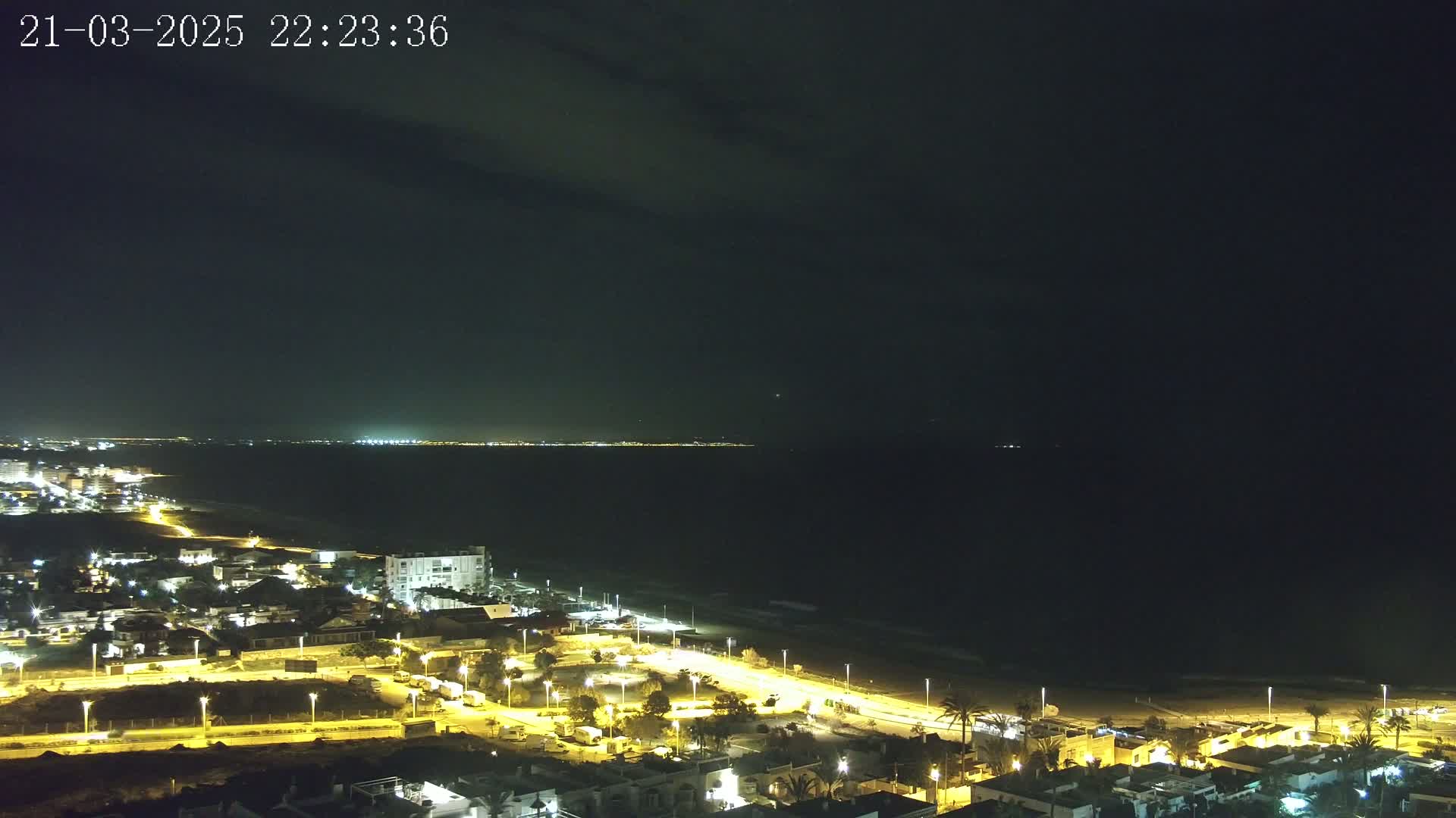 A nighttime coastal town illuminated by streetlights overlooks a dark ocean under a mostly clear sky.