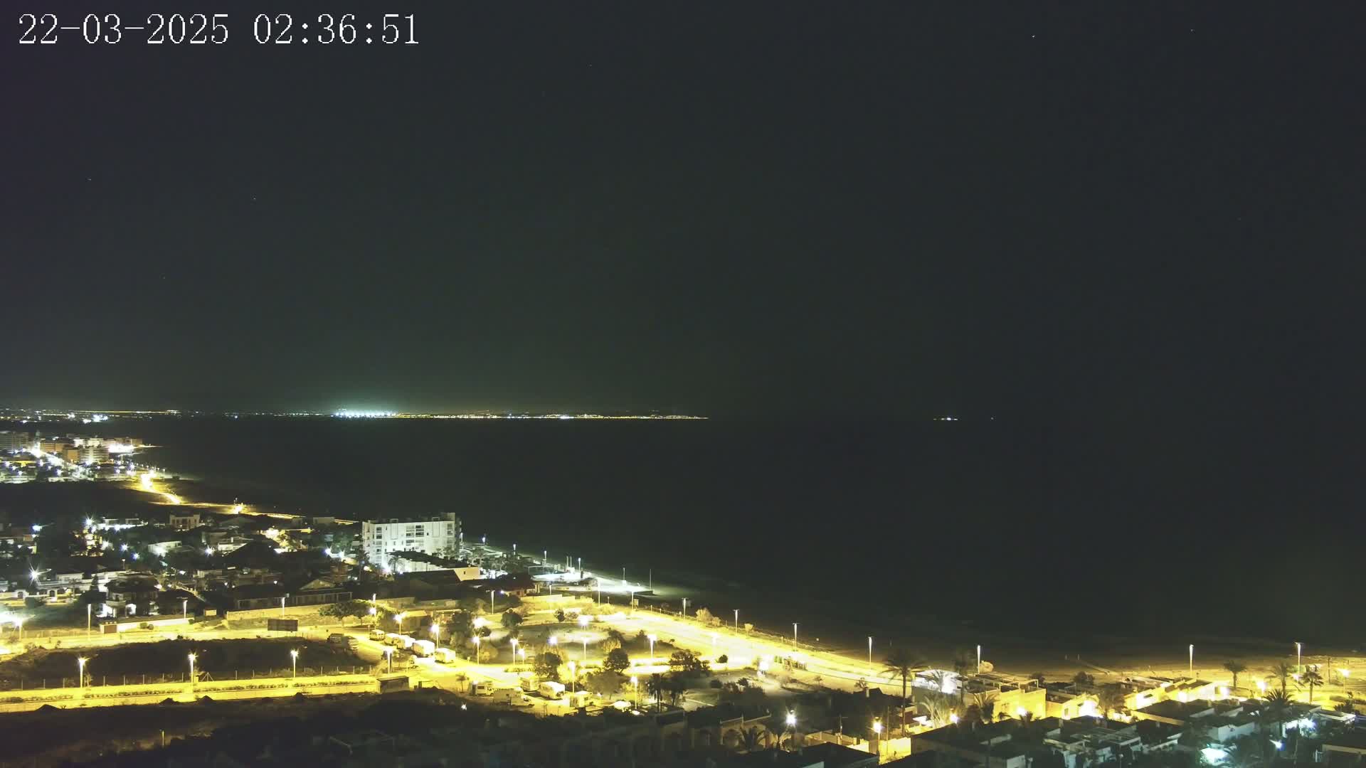 A nighttime coastal scene shows a dark ocean under a clear sky, with a town's illuminated streets and buildings extending along a sandy beach.