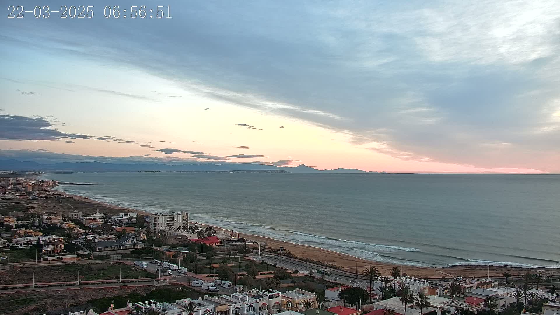An aerial view of a coastal town at sunset, showing a calm sea with gentle waves lapping a sandy beach, under a partly cloudy sky with pink and blue hues.