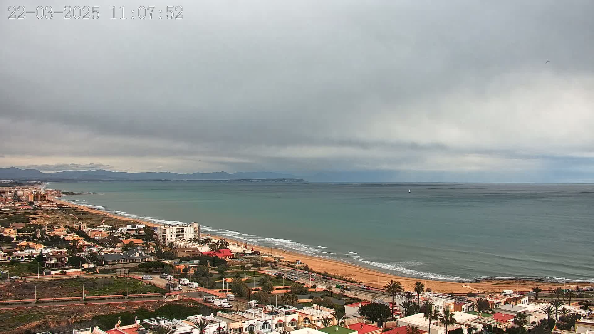 A coastal town under an overcast sky overlooks a long sandy beach and calm sea with distant mountains visible on the horizon.