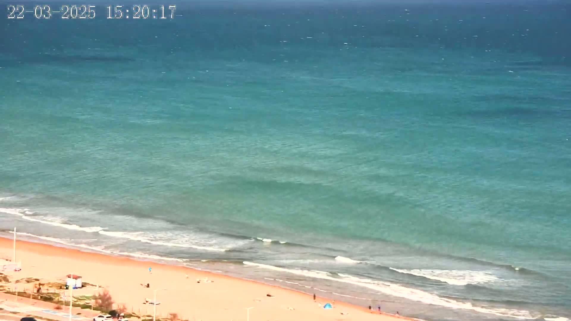 A sandy beach with a few people on it is gently lapped by turquoise water under a clear sky.