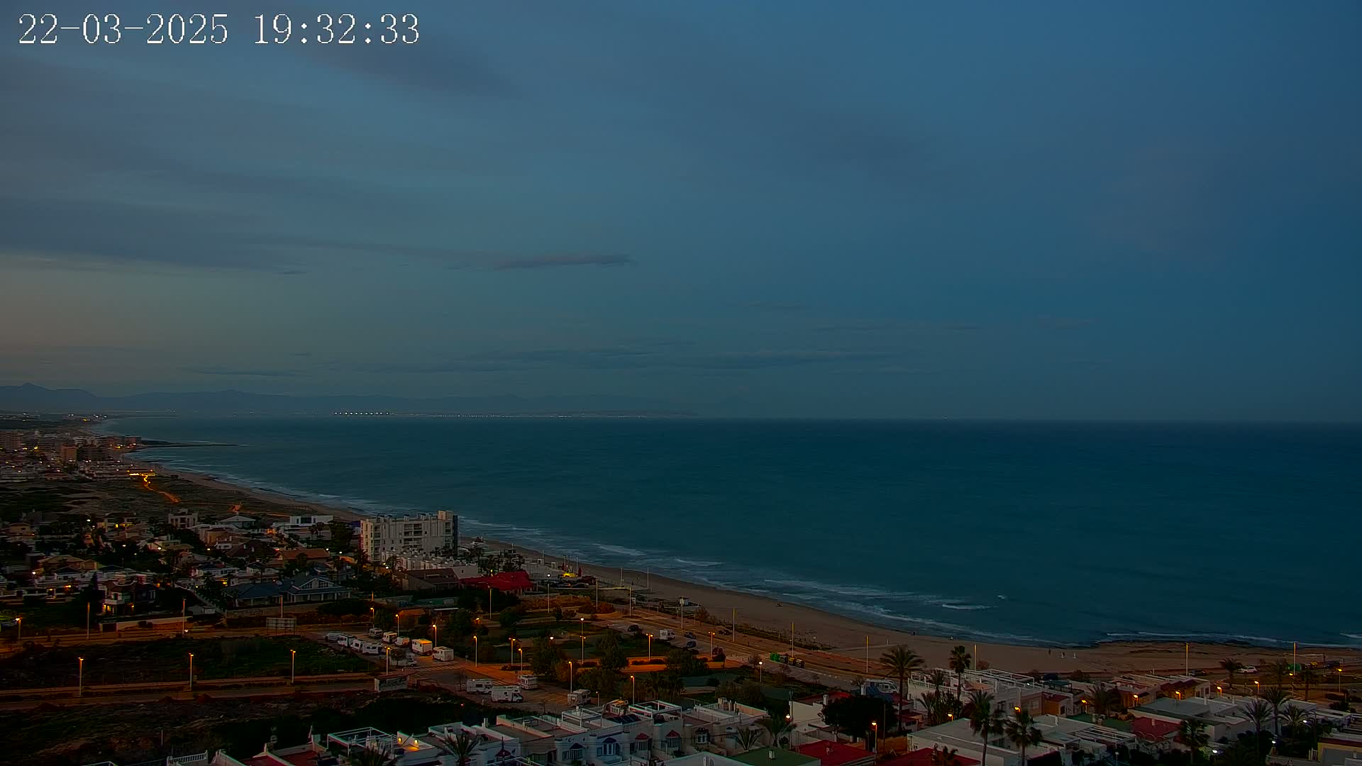 A coastal town at twilight is shown with its lights illuminated, overlooking a dark blue ocean under a mostly clear sky.