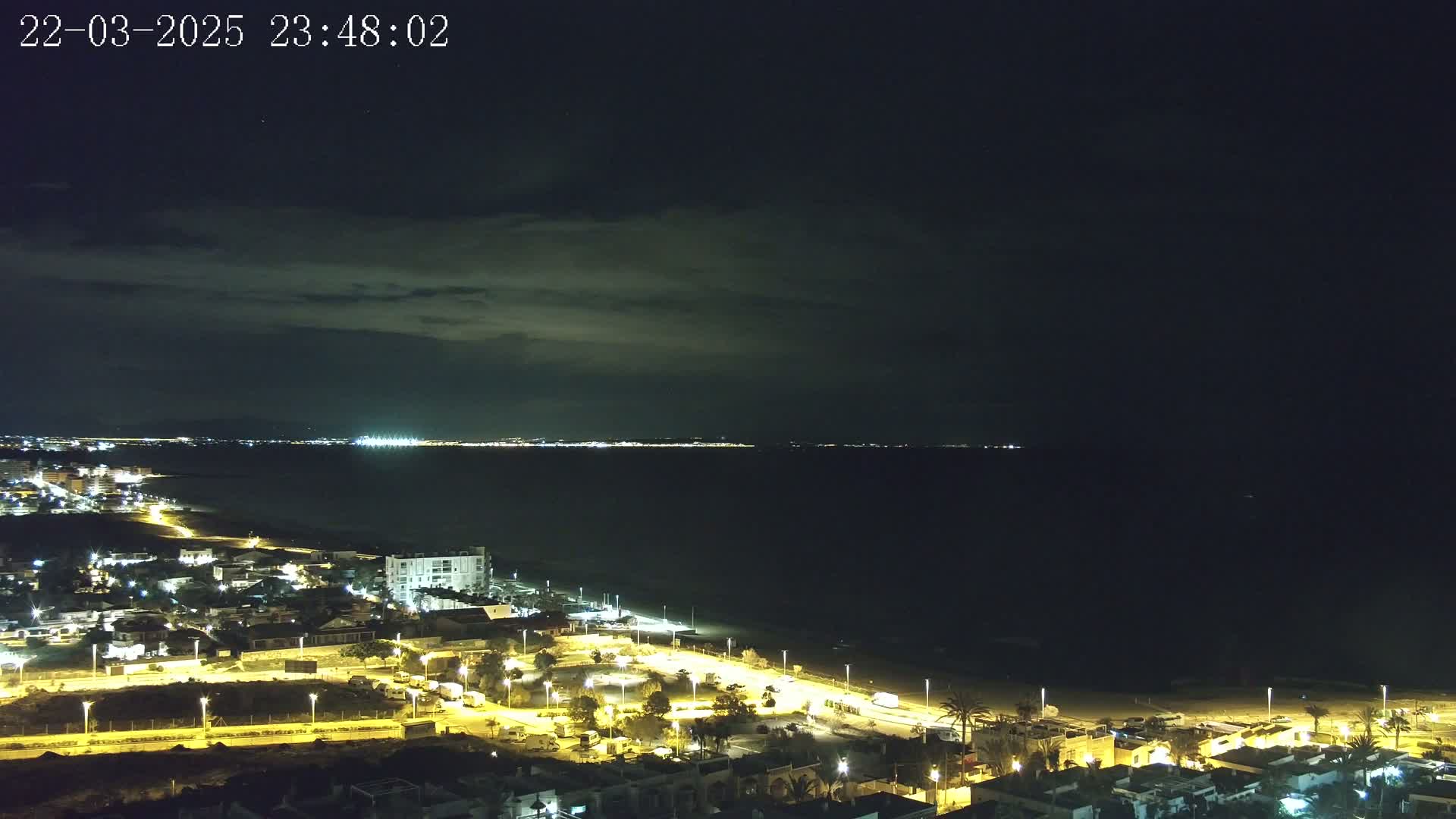 A nighttime coastal view shows a dark sea under a partly cloudy sky, with a brightly lit town visible along the shore and distant city lights across the water.