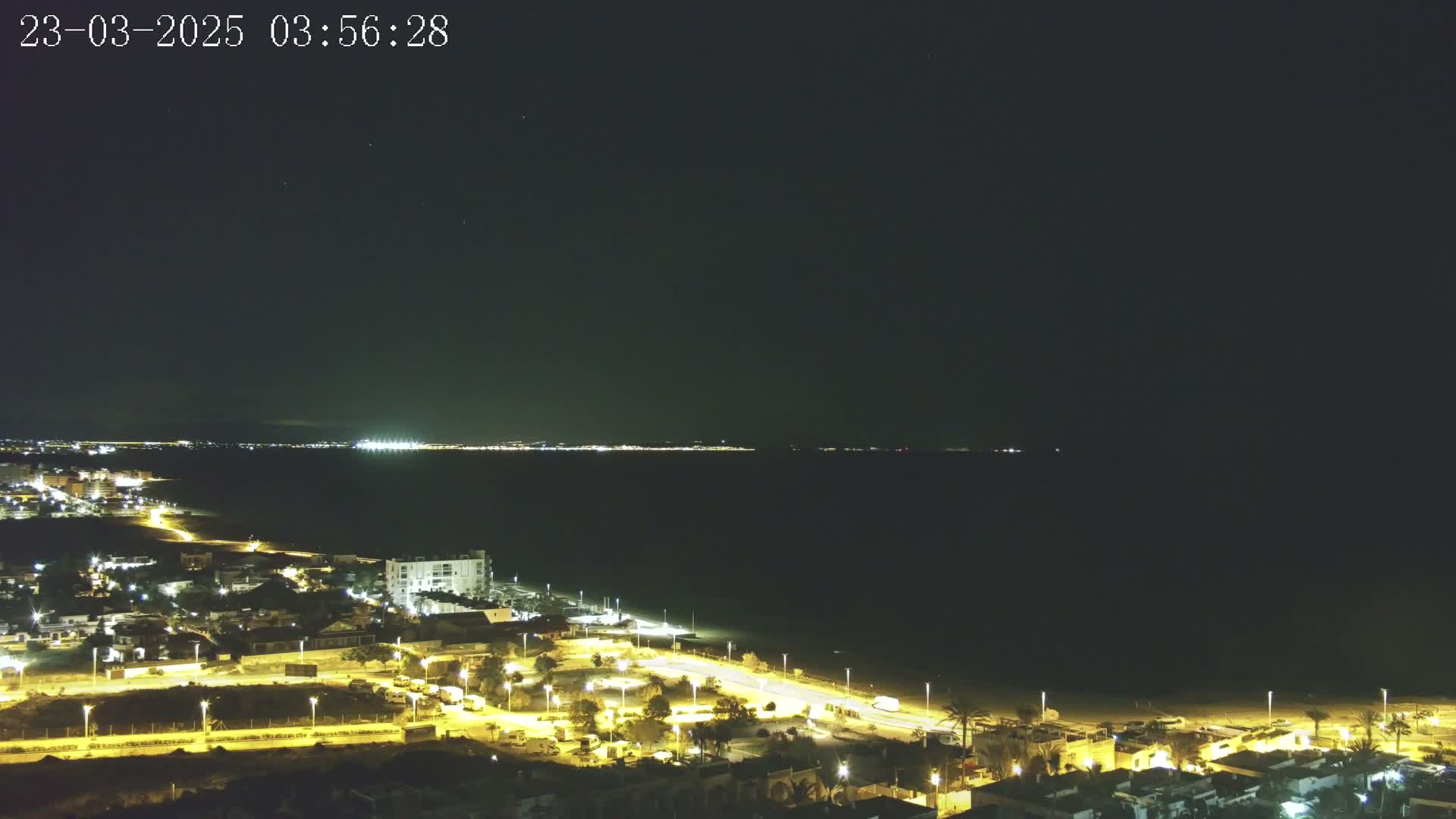 A nighttime coastal scene shows a dark sea with a brightly lit town on the shore under a clear, dark sky.