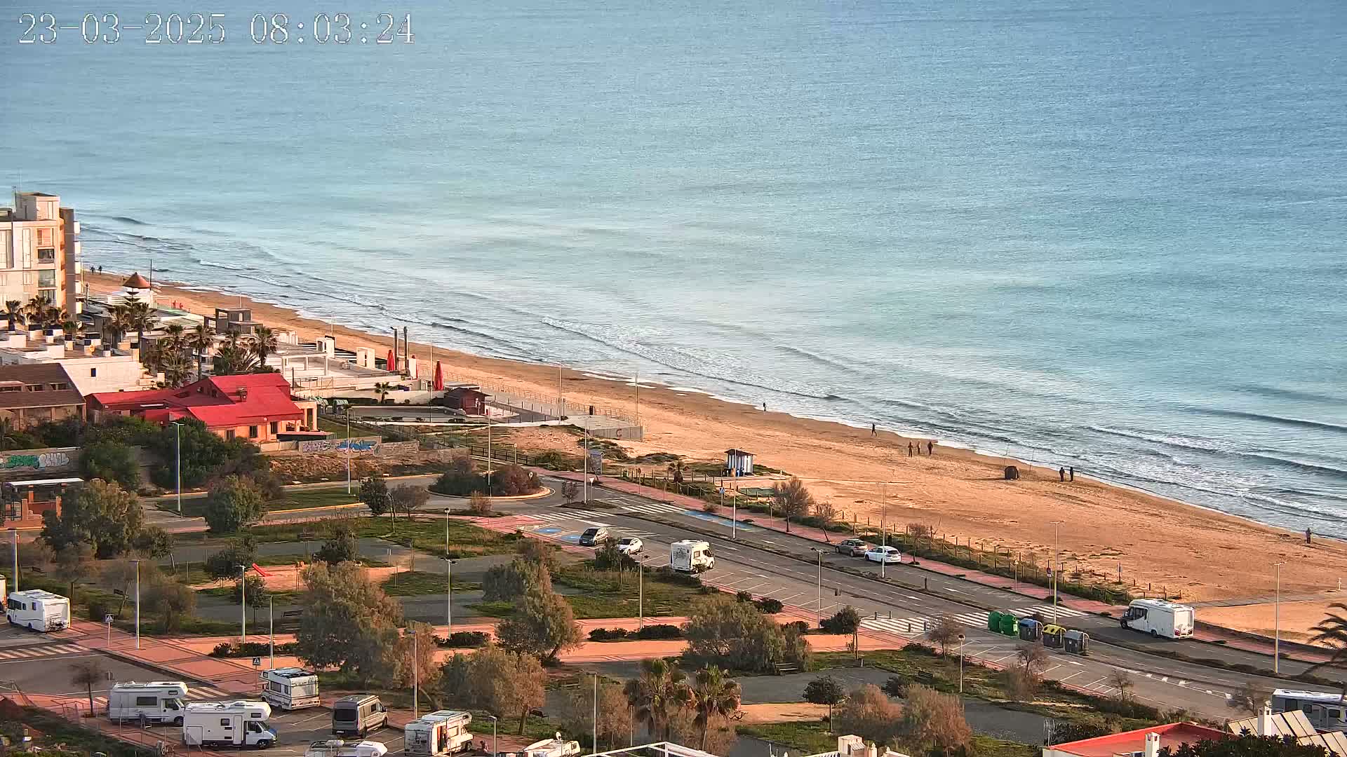 A sandy beach with gentle waves under a clear sky, bordered by a road with several recreational vehicles parked nearby, and buildings and vegetation in the background.