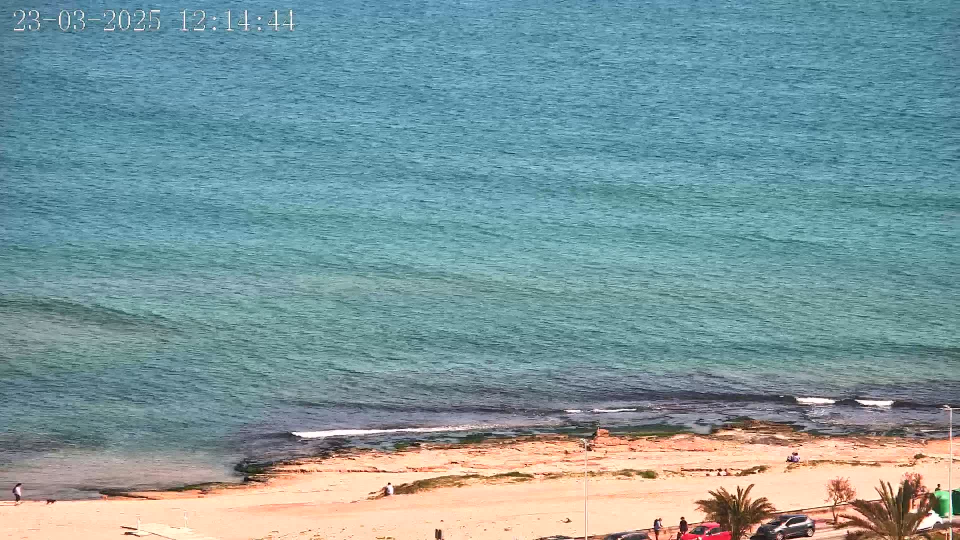 A sandy beach with a few people on it borders a calm, teal ocean under a sunny sky.