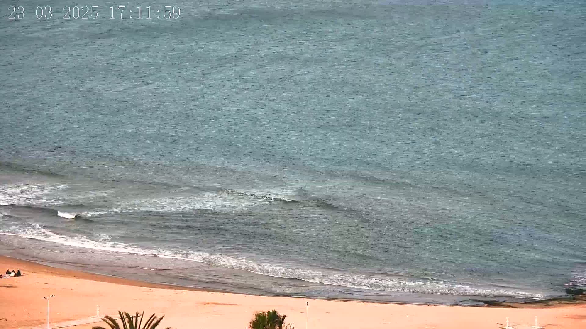 An overhead view shows a sandy beach meeting a calm, teal ocean with small waves under an overcast sky.
