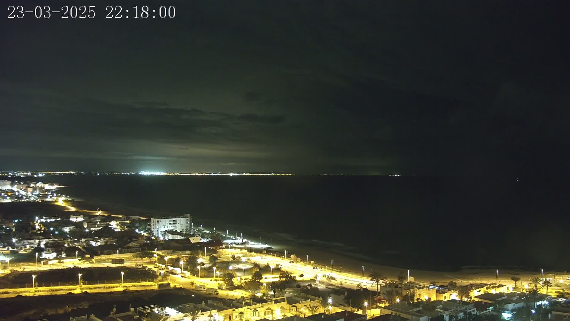 A nighttime coastal scene shows a dark sea under a cloudy sky, with a brightly lit town stretching along a sandy beach.