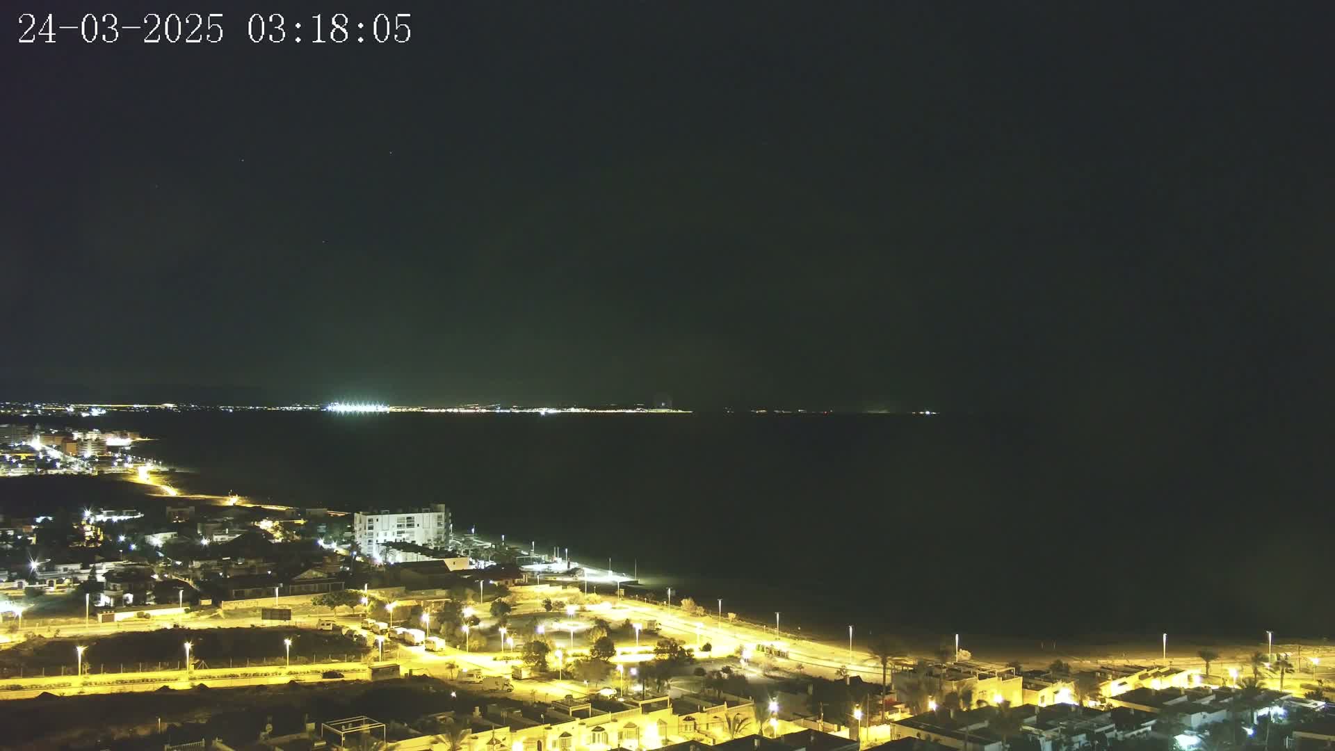 A coastal town at night, illuminated by streetlights, overlooks a dark ocean under a clear, starlit sky.