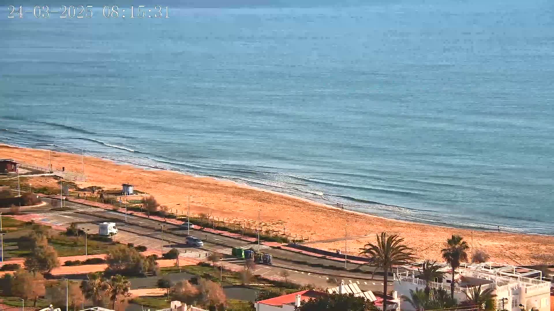 An aerial view shows a sandy beach meeting a calm, blue ocean under a sunny sky, with a road and buildings visible beyond the beach.