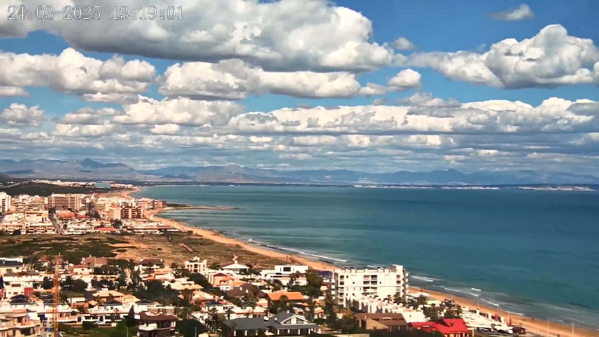 A coastal town with a sandy beach and blue ocean stretches out under a partly cloudy sky.