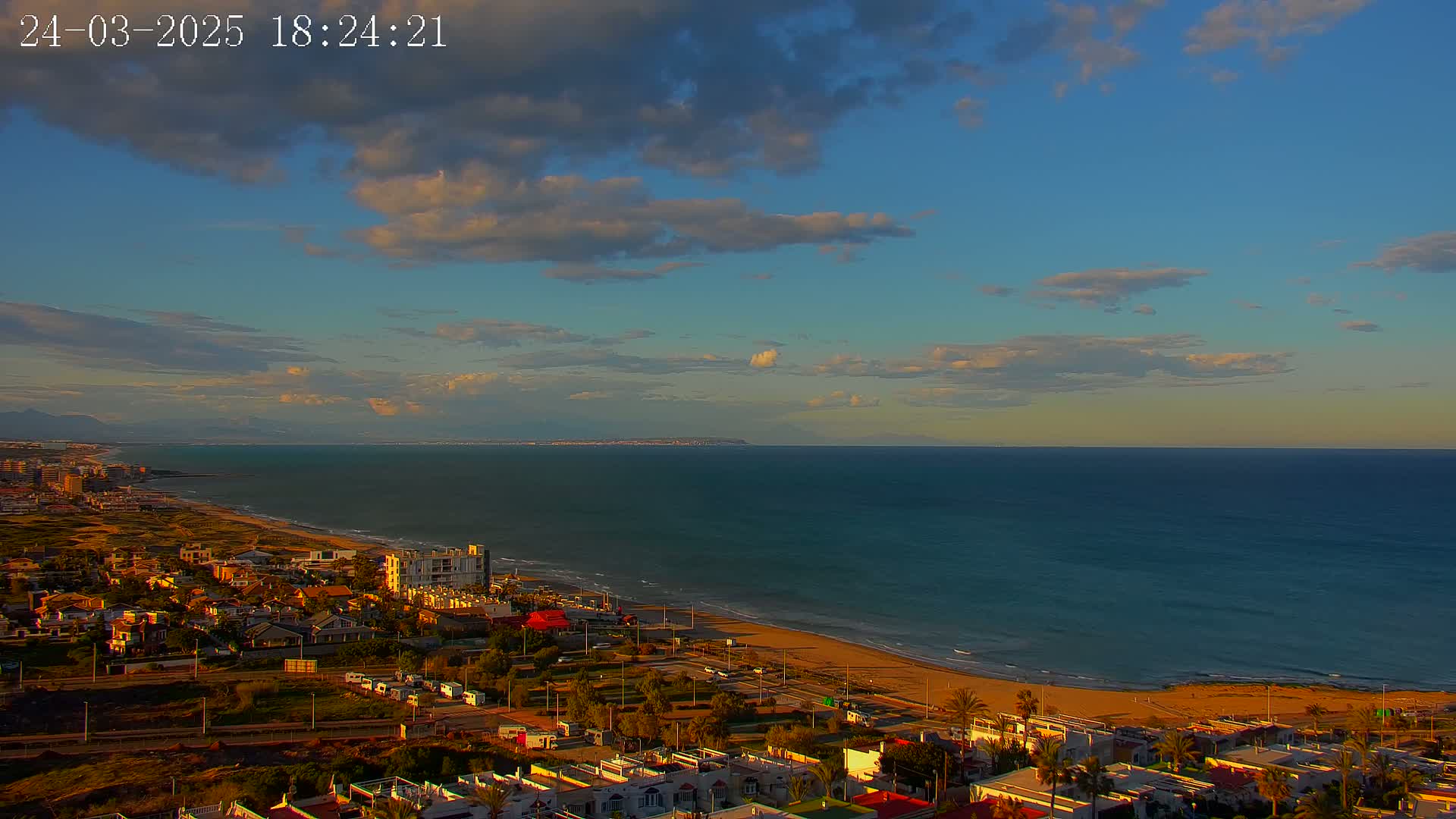 A coastal town with a sandy beach and calm ocean under a partly cloudy sunset sky.