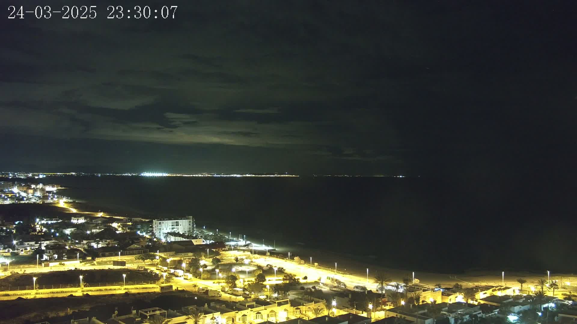 A nighttime coastal scene shows a dark sea under a partly cloudy sky, with a brightly lit town and beach visible in the foreground.