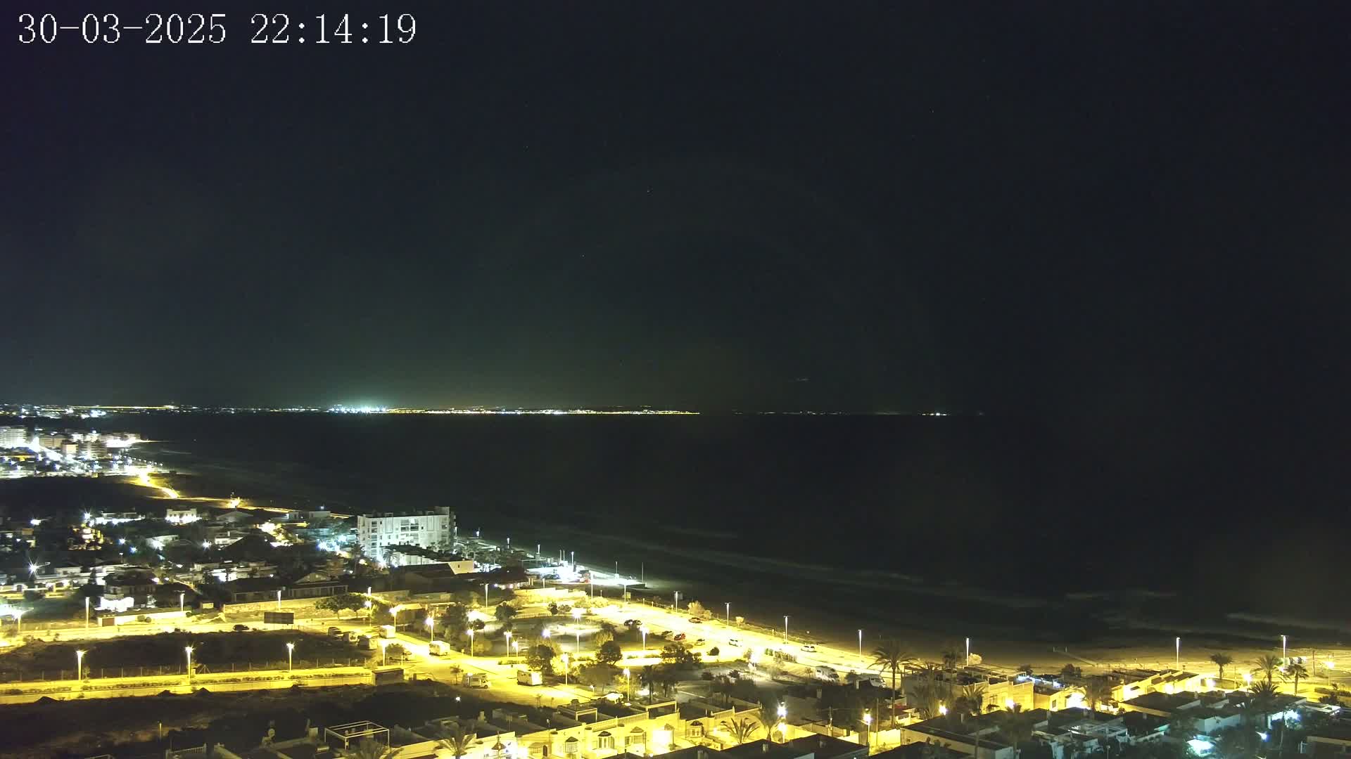 A nighttime view of a coastal town illuminated by streetlights, overlooking a dark ocean with a faint horizon line of distant city lights under a clear, dark sky.