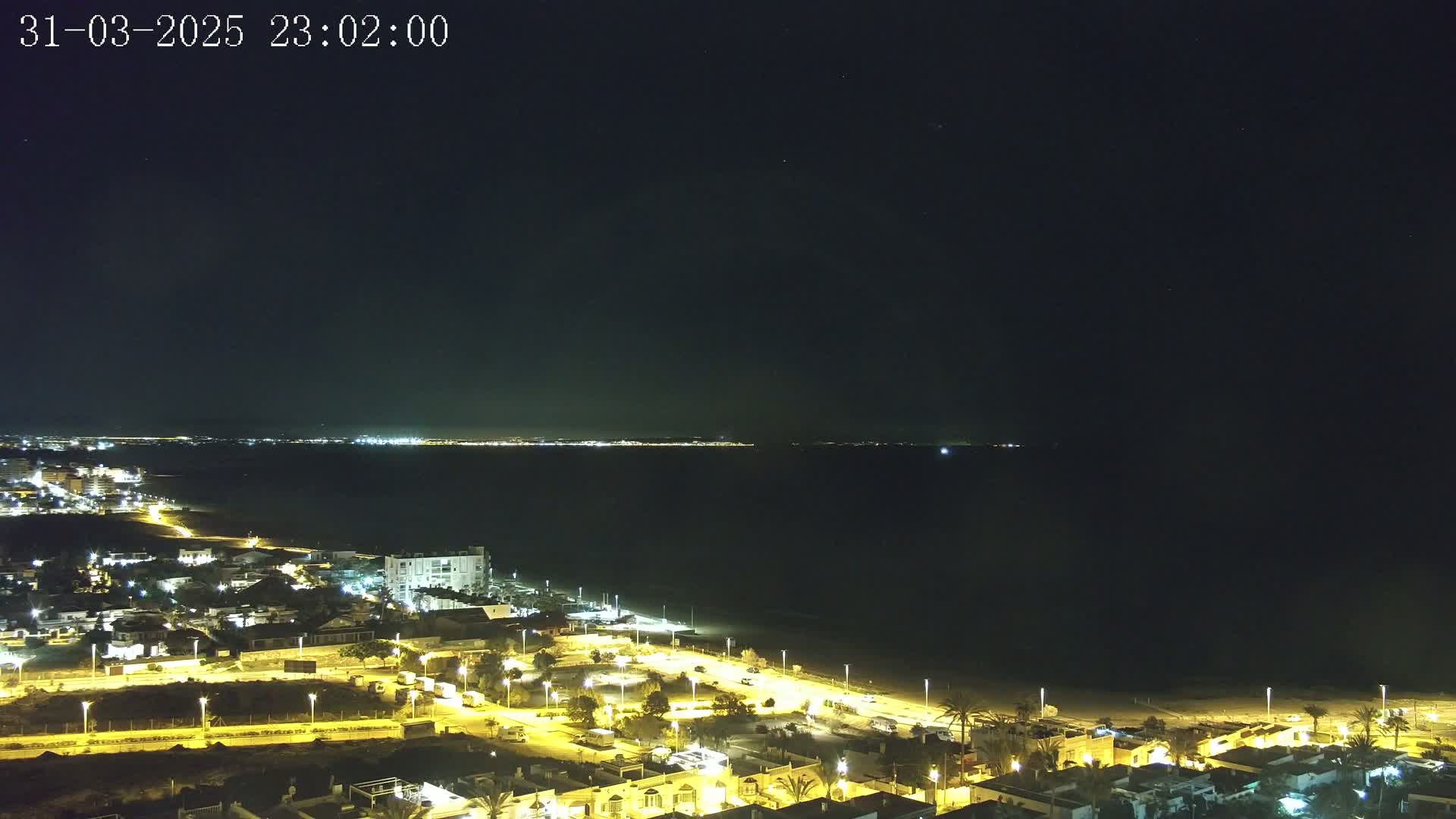 A nighttime coastal view shows a dark sea and a brightly lit town with streetlights, under a clear, starry sky.