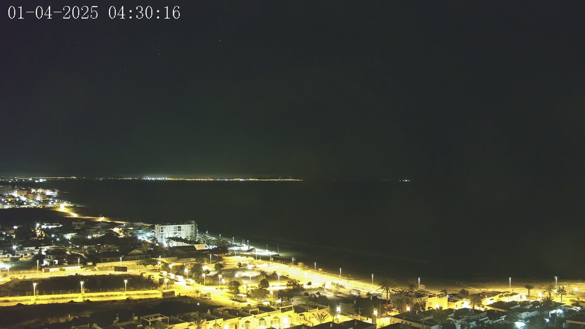 A nighttime coastal scene shows a dark ocean, a lit beach, and a town with illuminated streets and buildings under a clear, dark sky.