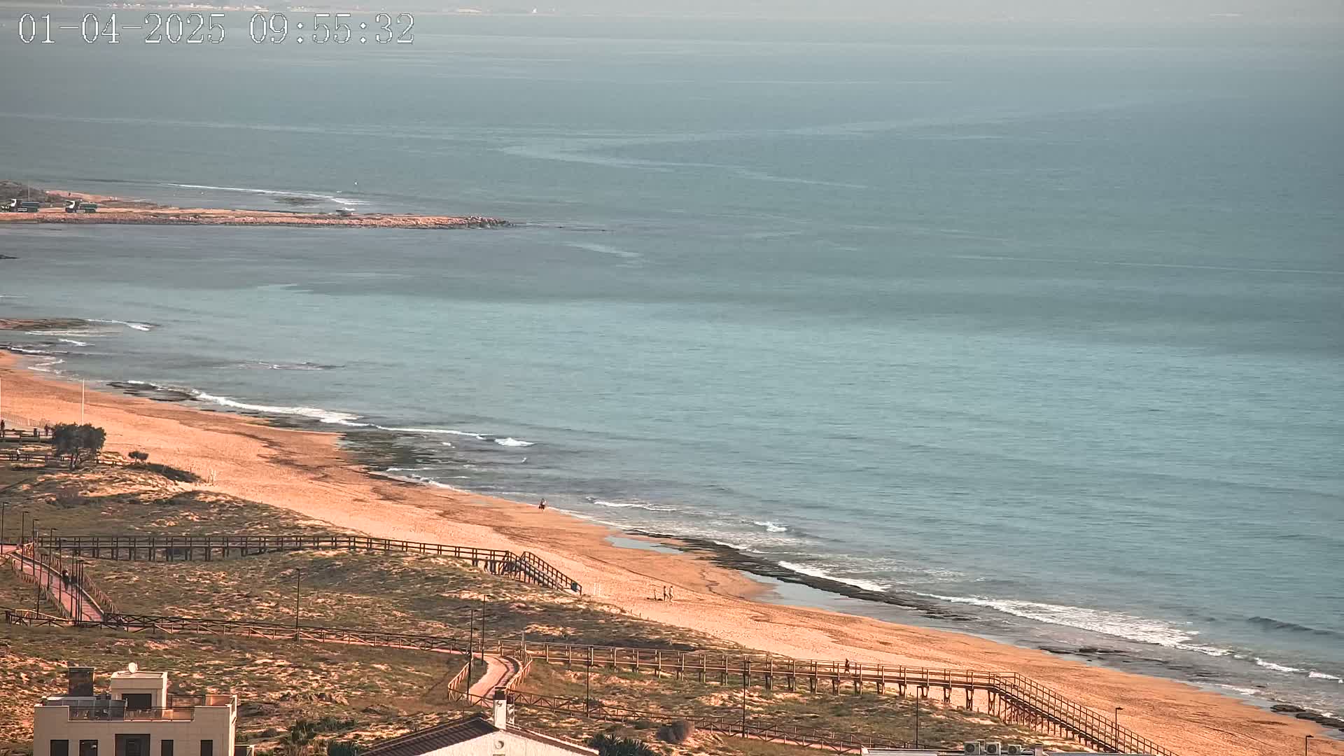 A sandy beach with a wooden boardwalk stretches along a calm, teal ocean under a hazy sky.