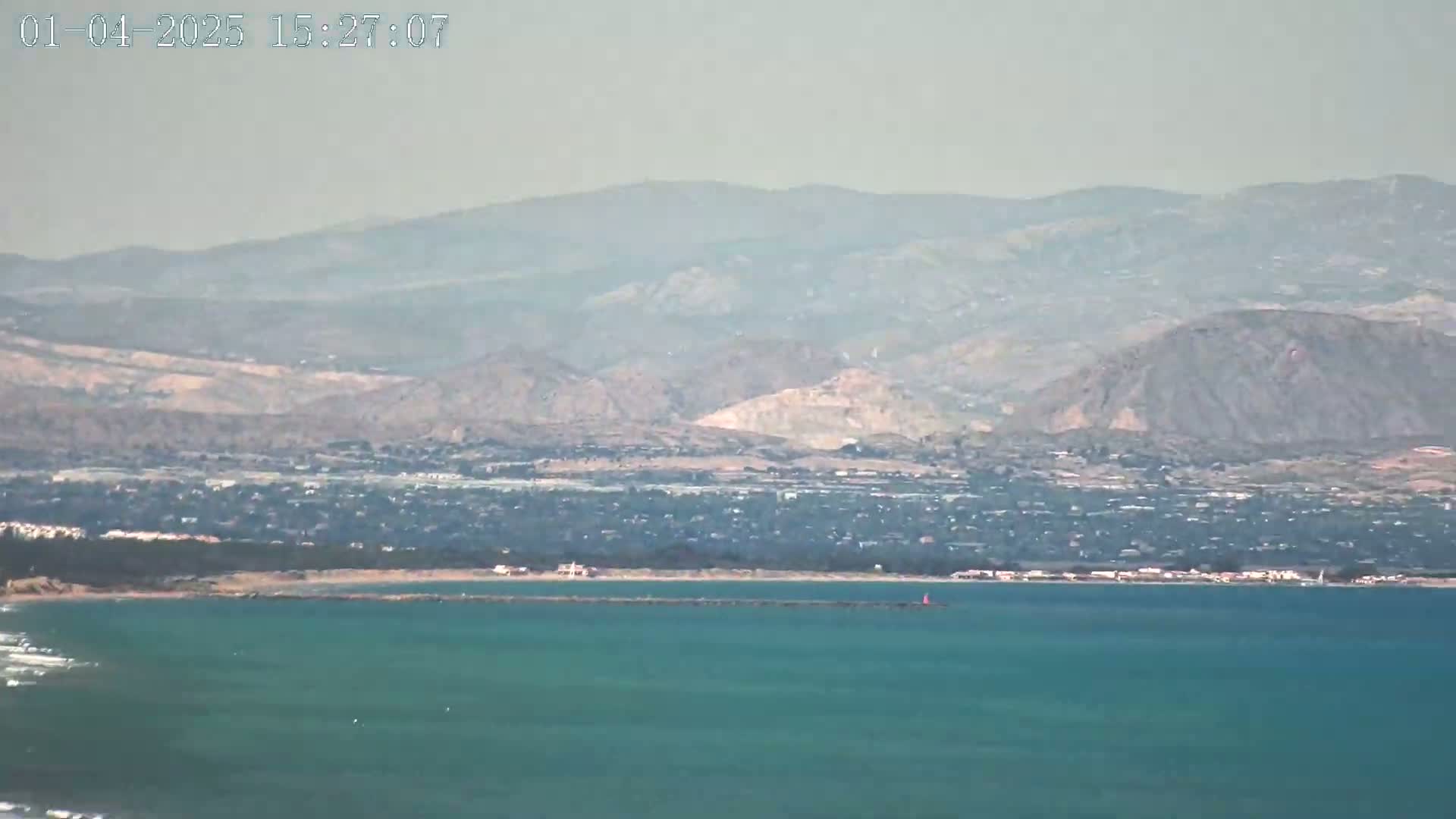 A hazy view shows a teal ocean, a coastline with a town, and distant brown mountains under a clear sky.