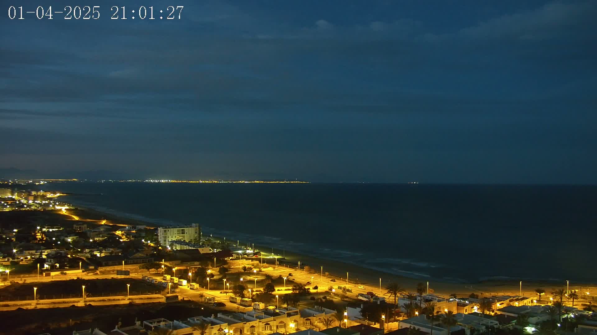 A nighttime coastal town with illuminated buildings and streets stretches along a sandy beach, overlooking a dark, calm sea under a mostly clear, dusky blue sky.