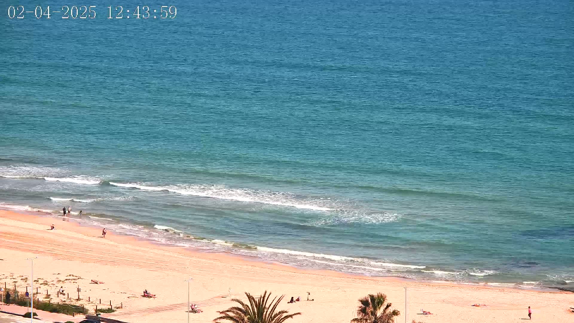 A sandy beach with a few people on it meets a calm, teal ocean with small waves under a sunny sky.