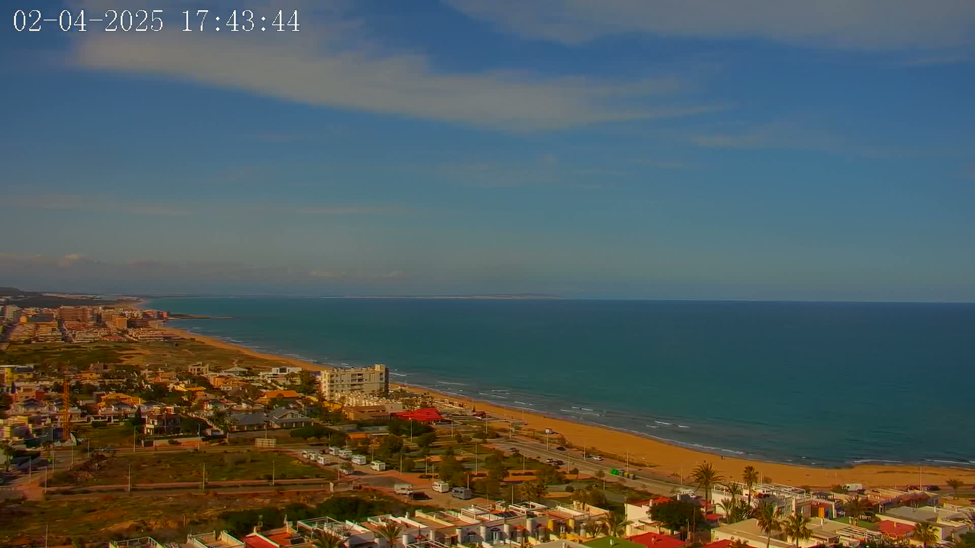 An aerial view shows a coastal town on a sunny day with a mostly clear sky and calm ocean waves rolling onto a sandy beach.