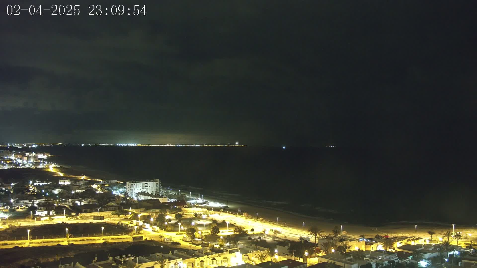 A nighttime coastal town is illuminated, with a dark, cloudy sky above and a calm, dark ocean in the distance.