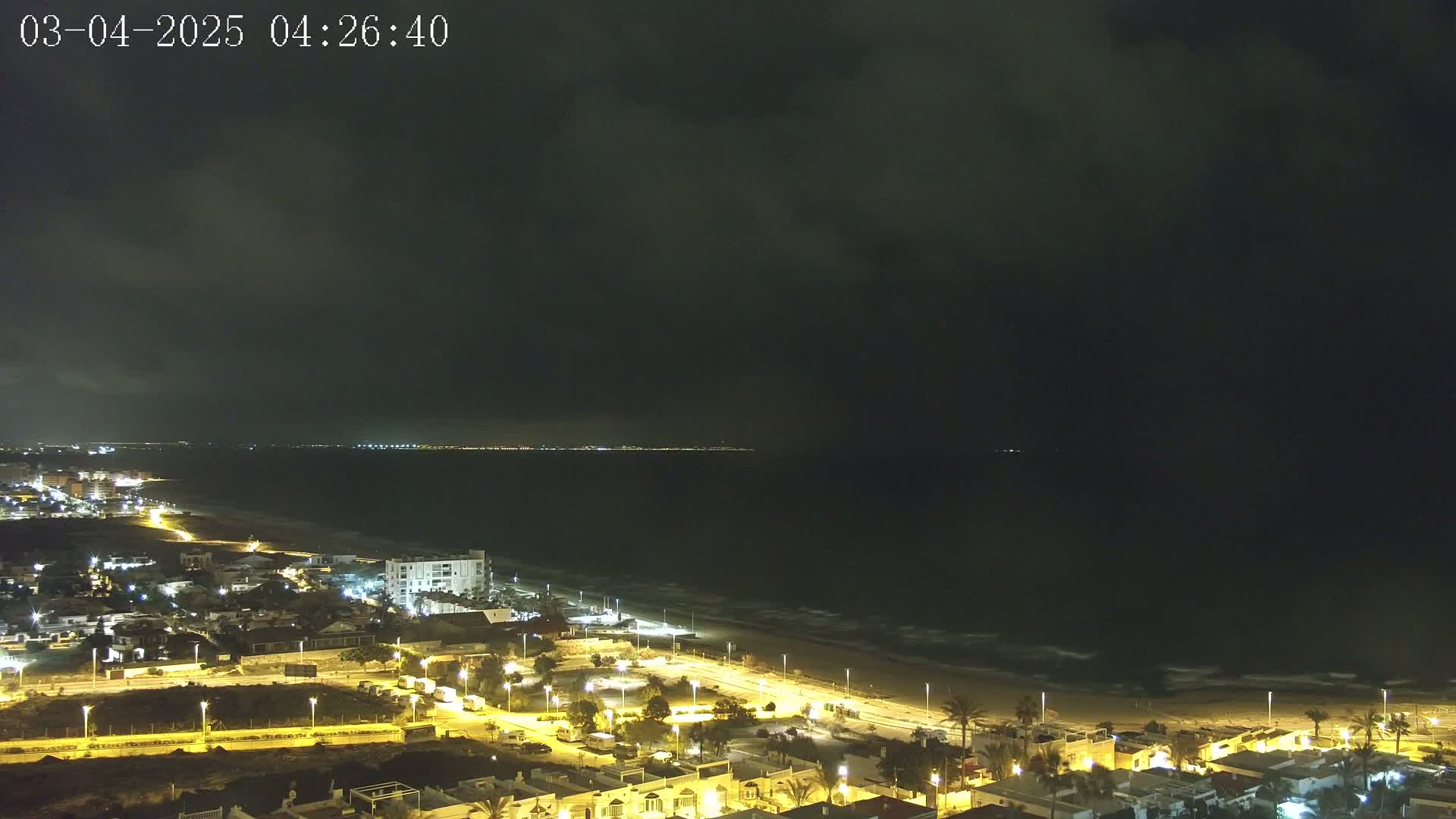 A nighttime coastal town with illuminated streets and buildings is shown, overlooking a dark ocean with mild waves under a cloudy sky.