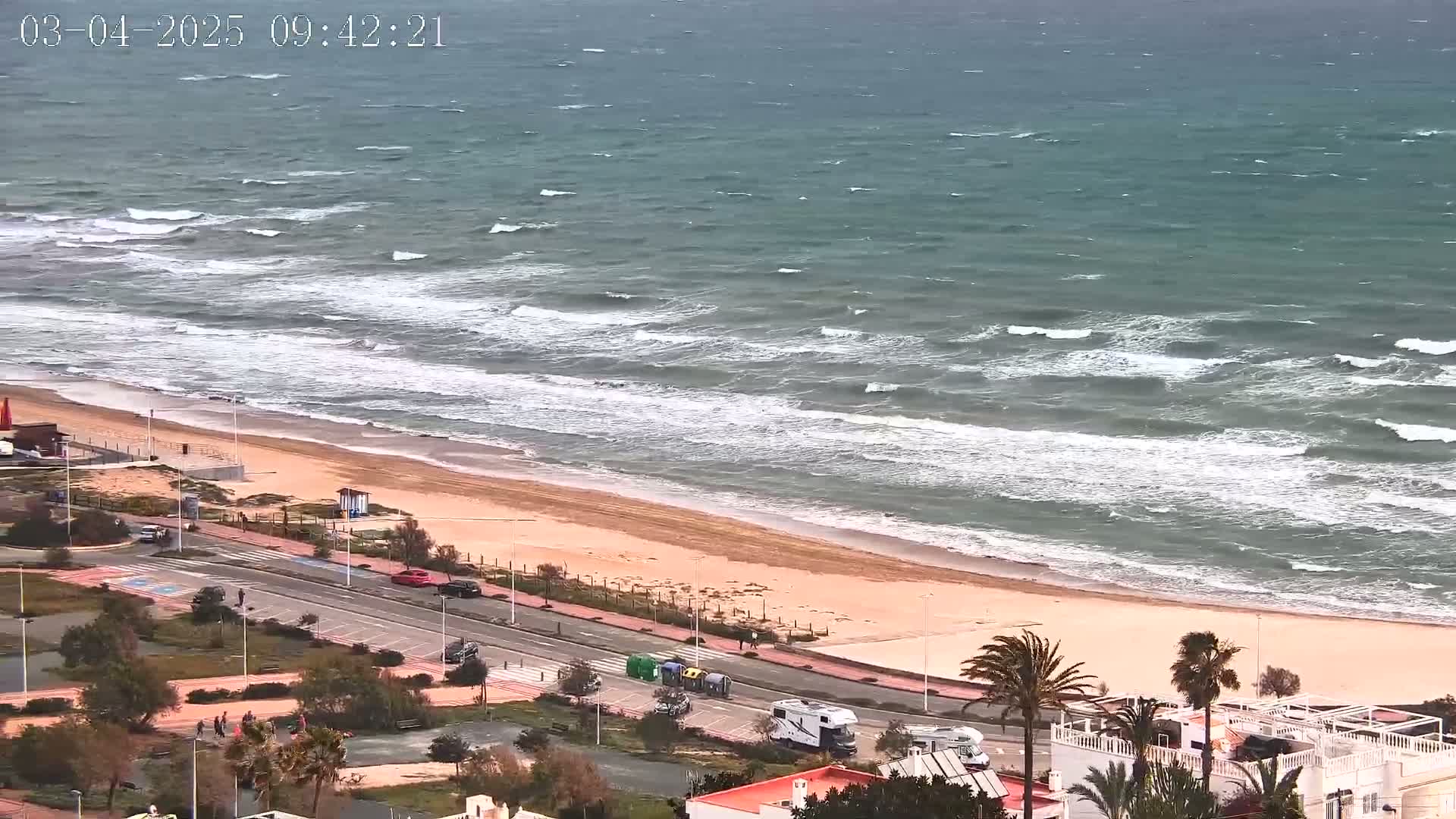 An aerial view shows a beach with choppy waves under a cloudy sky, and a road with cars and buildings beyond.