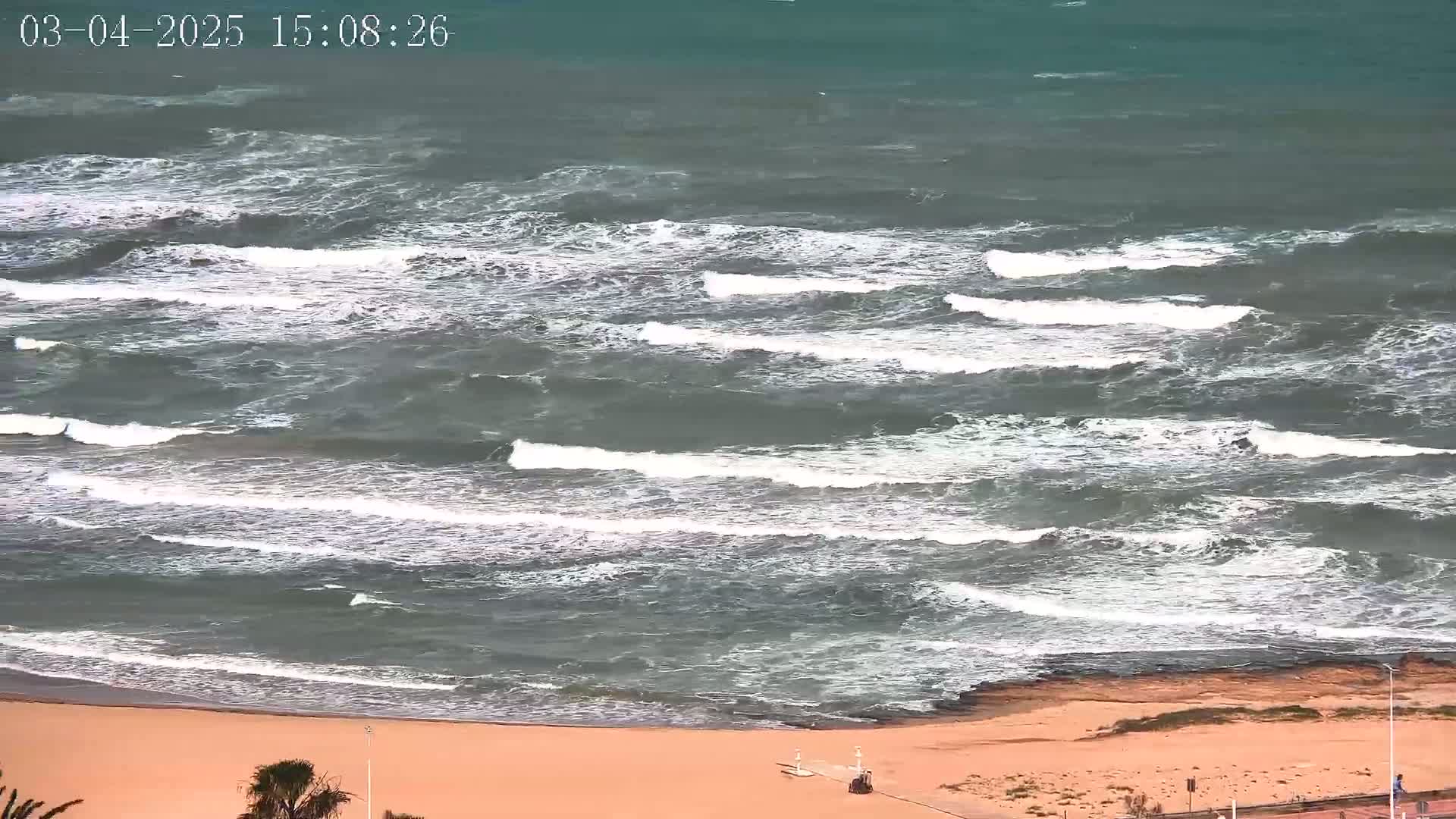 A sandy beach with moderately sized waves crashing on the shore under somewhat overcast skies.