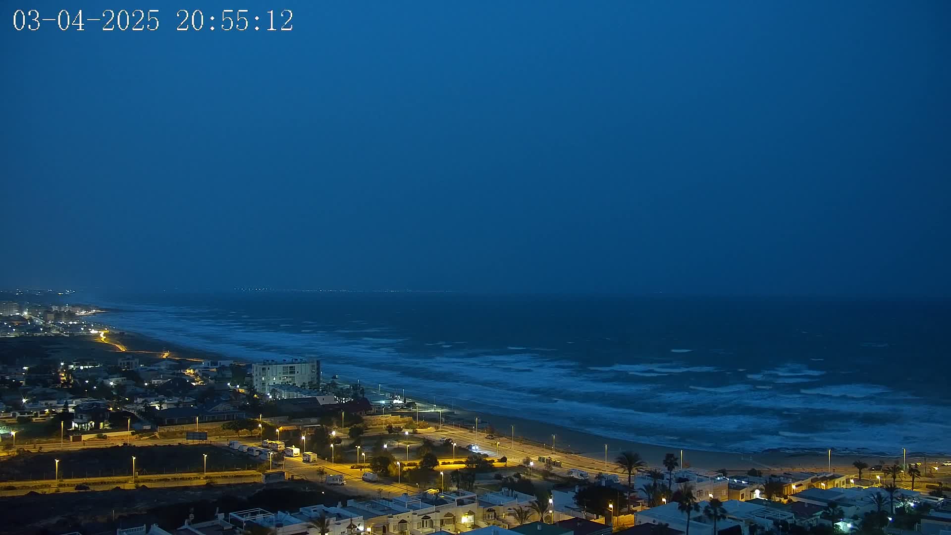 A nighttime coastal scene shows a dark blue sky over a wavy ocean, with a town's lights illuminating the beach and shoreline.