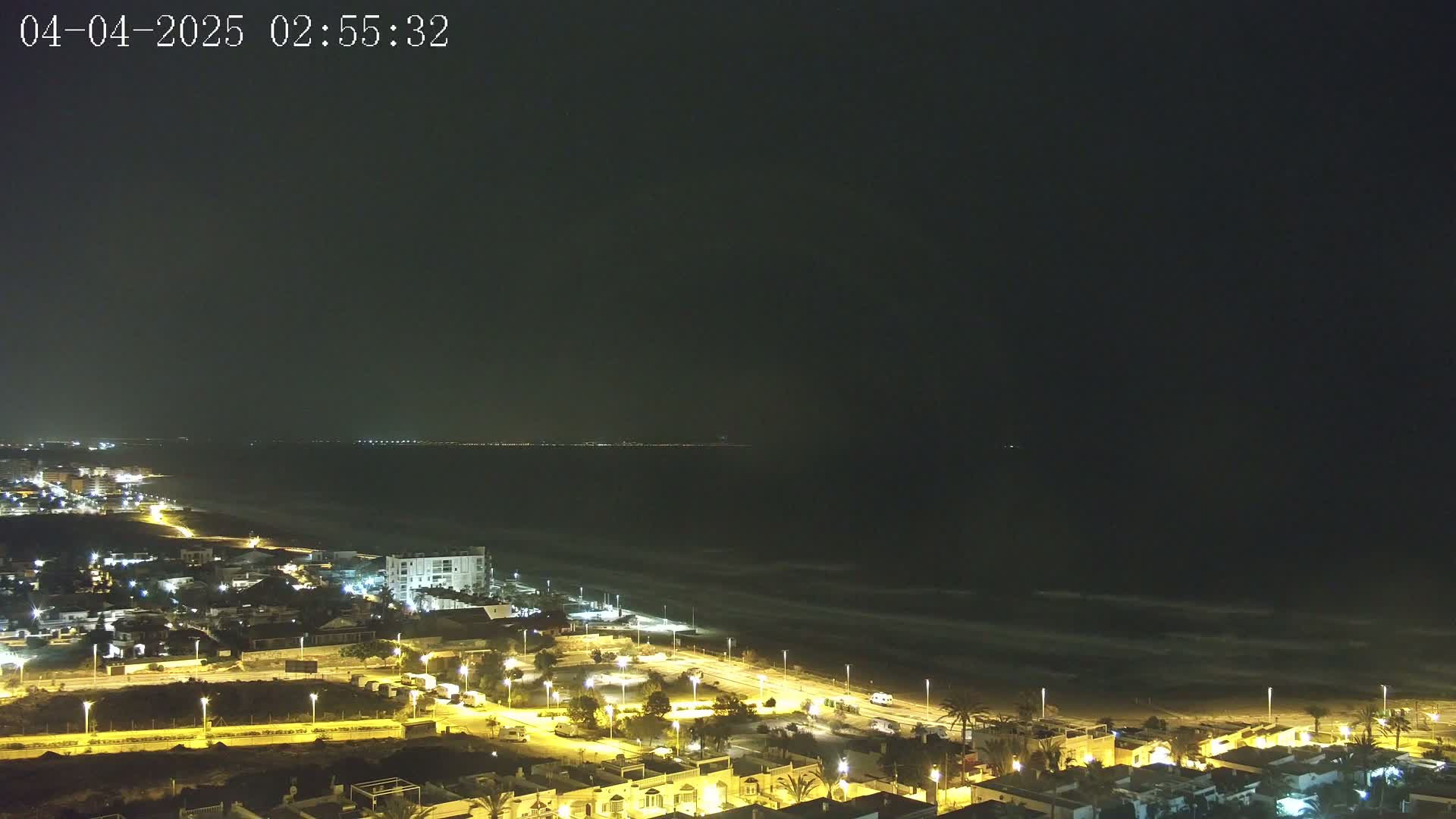 A nighttime coastal town with illuminated streets and buildings overlooks a dark ocean with gently lapping waves under a clear, dark sky.