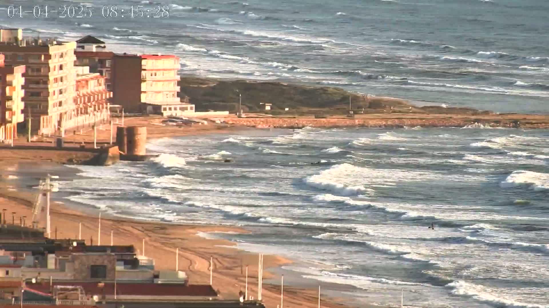 A sandy beach with moderate waves and several multi-story buildings is visible from an elevated vantage point.